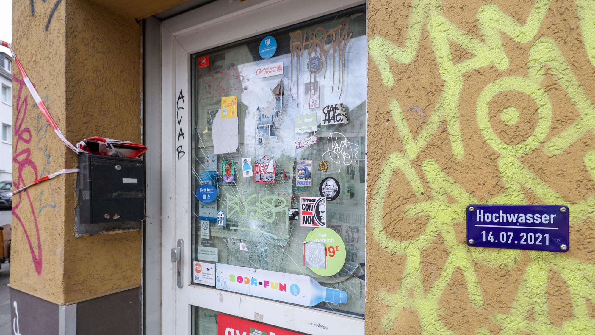 Eine Tür zu einem Kiosk ist zu sehen. Das Haus ist besprüht mit Graffiti. Eine Hochwassermarke ist an der Hauswand angebracht.