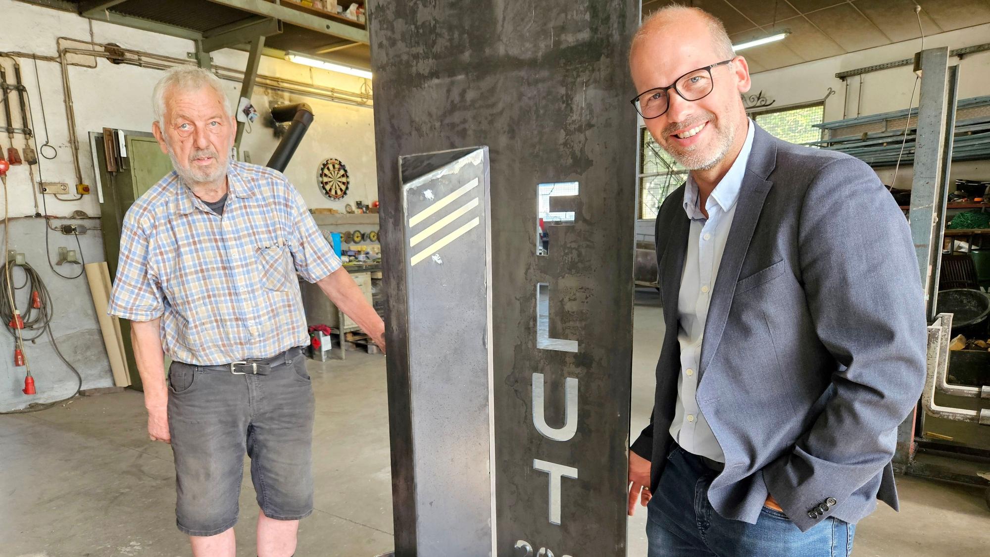 Zwei Männer stehen neben einer Metallstele, in die „Flut 2021“ eingraviert ist.