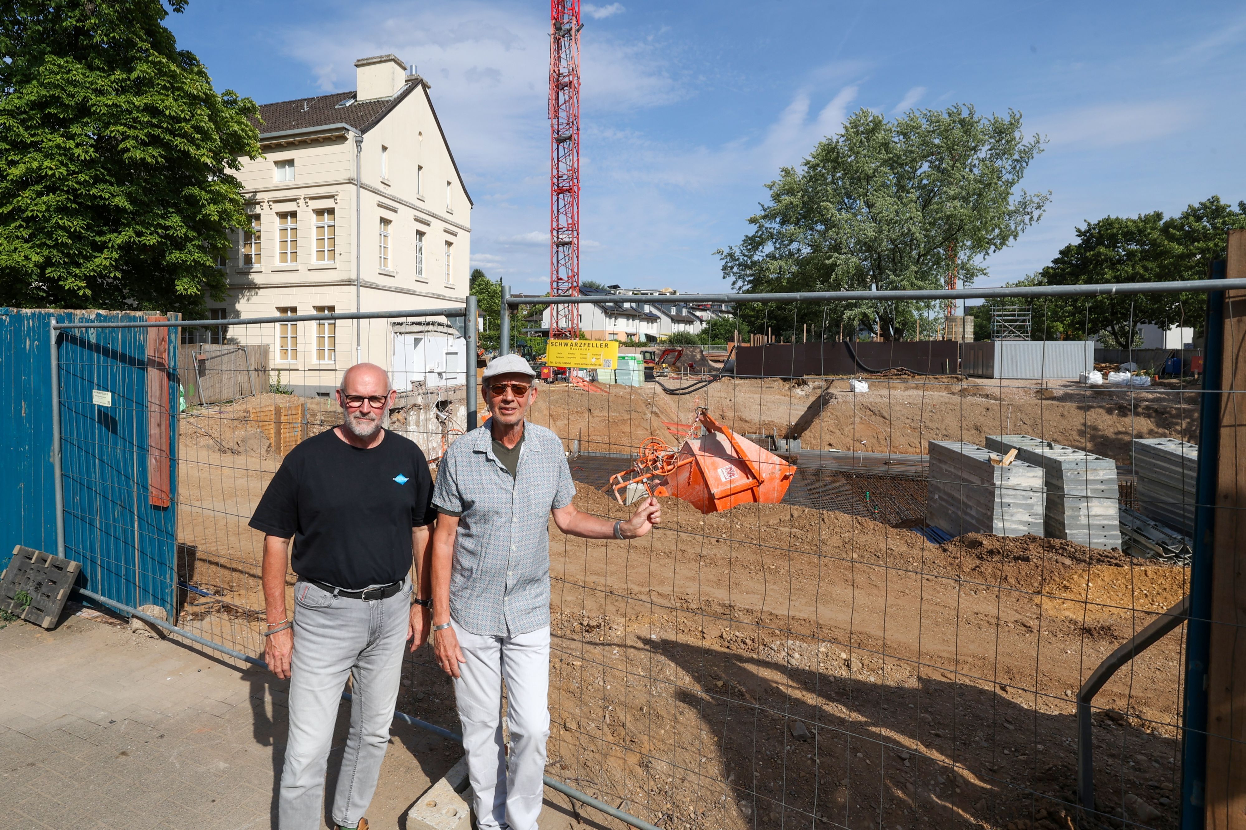 Zwei Männer stehen nebeneinander vor einem Bauzaun. Dahinter sind die Baustelle, ein Kran, und ein Gutshaus zu sehen.