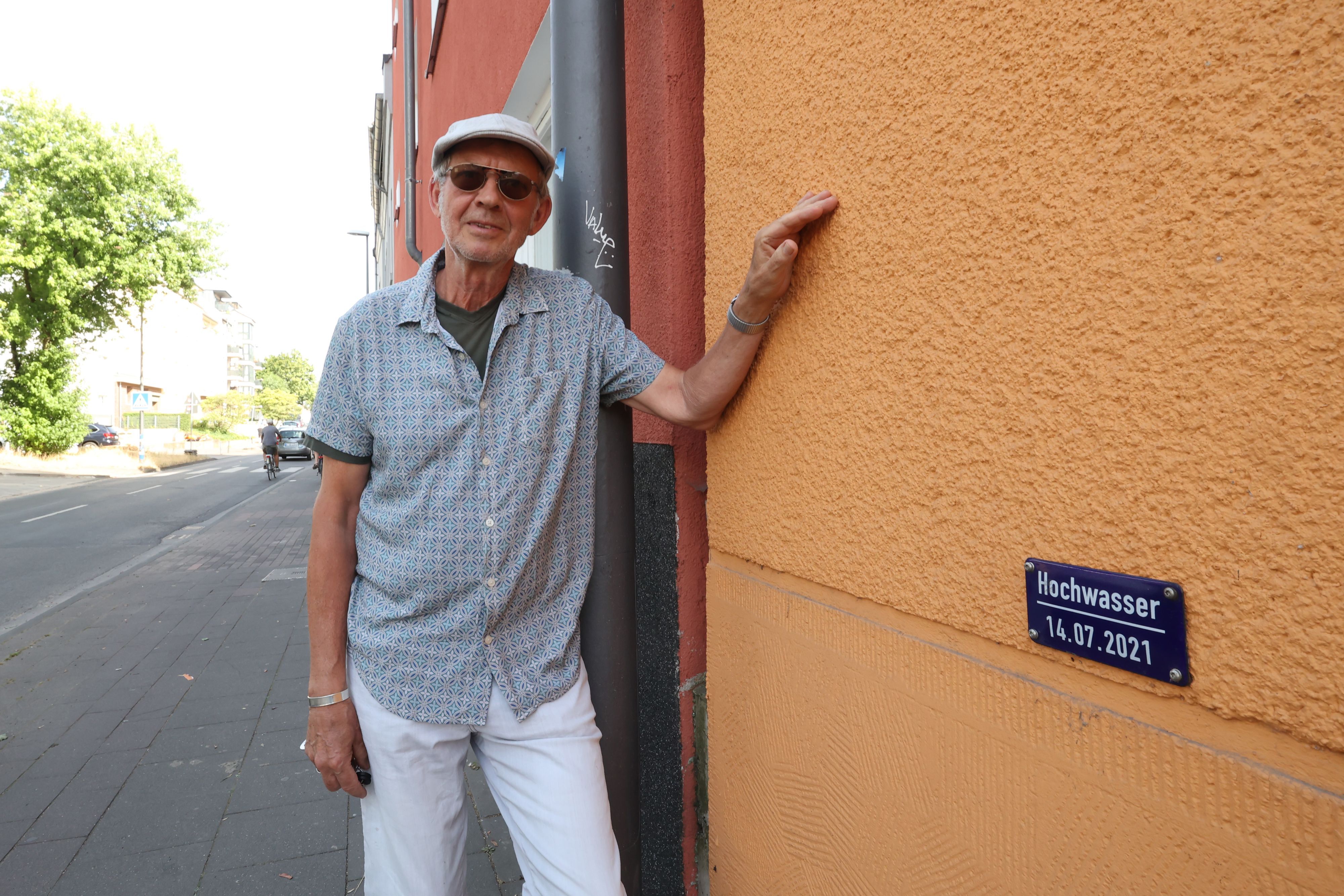 Ein Mann mit Kappe und Sonnenbrille steht vor einer Hauswand. Auf dieser ist eine Hochwassermarke angebracht.