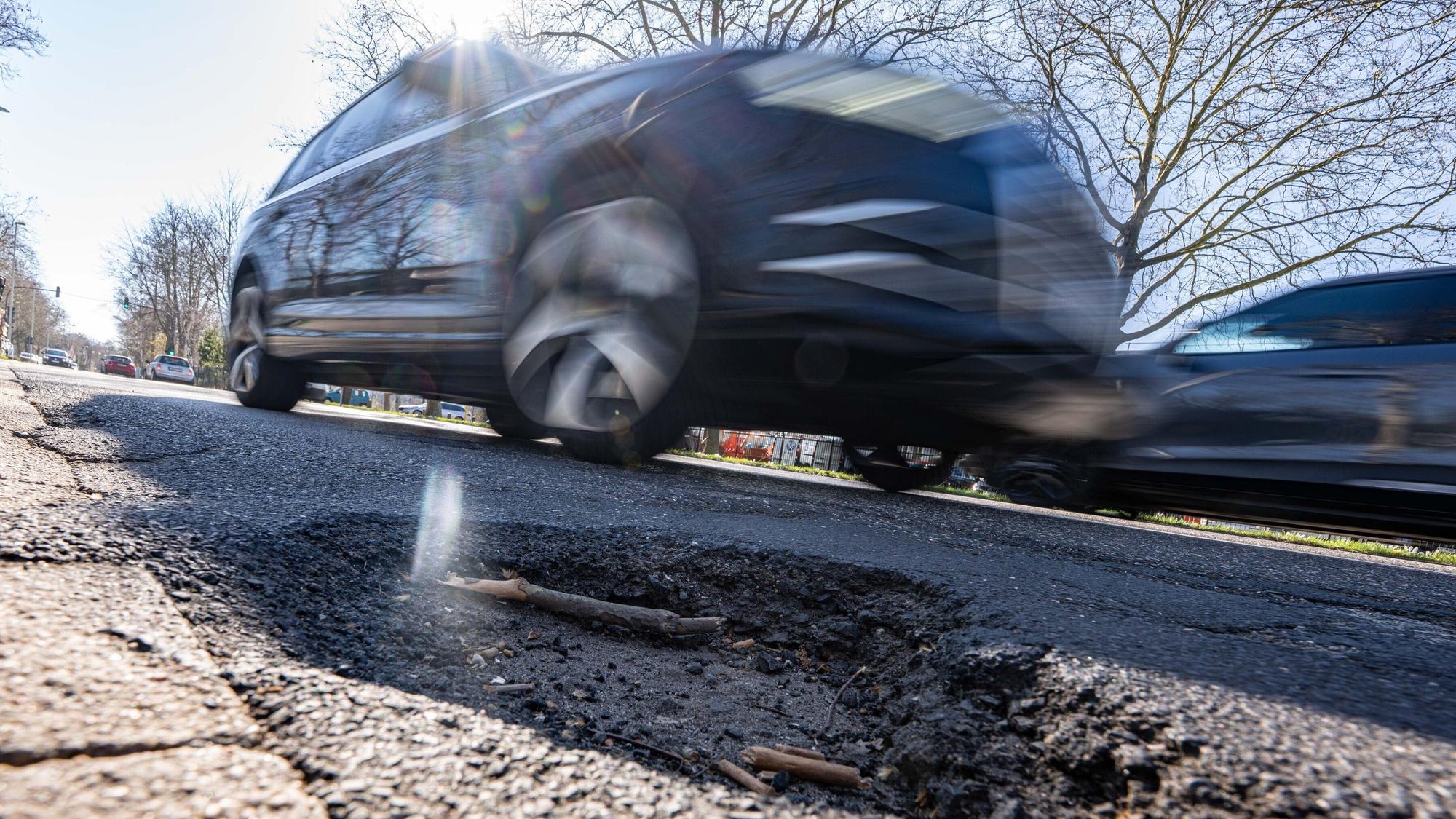 Das Foto zeigt ein Schlagloch, dahinter fährt ein Auto über die Straße.