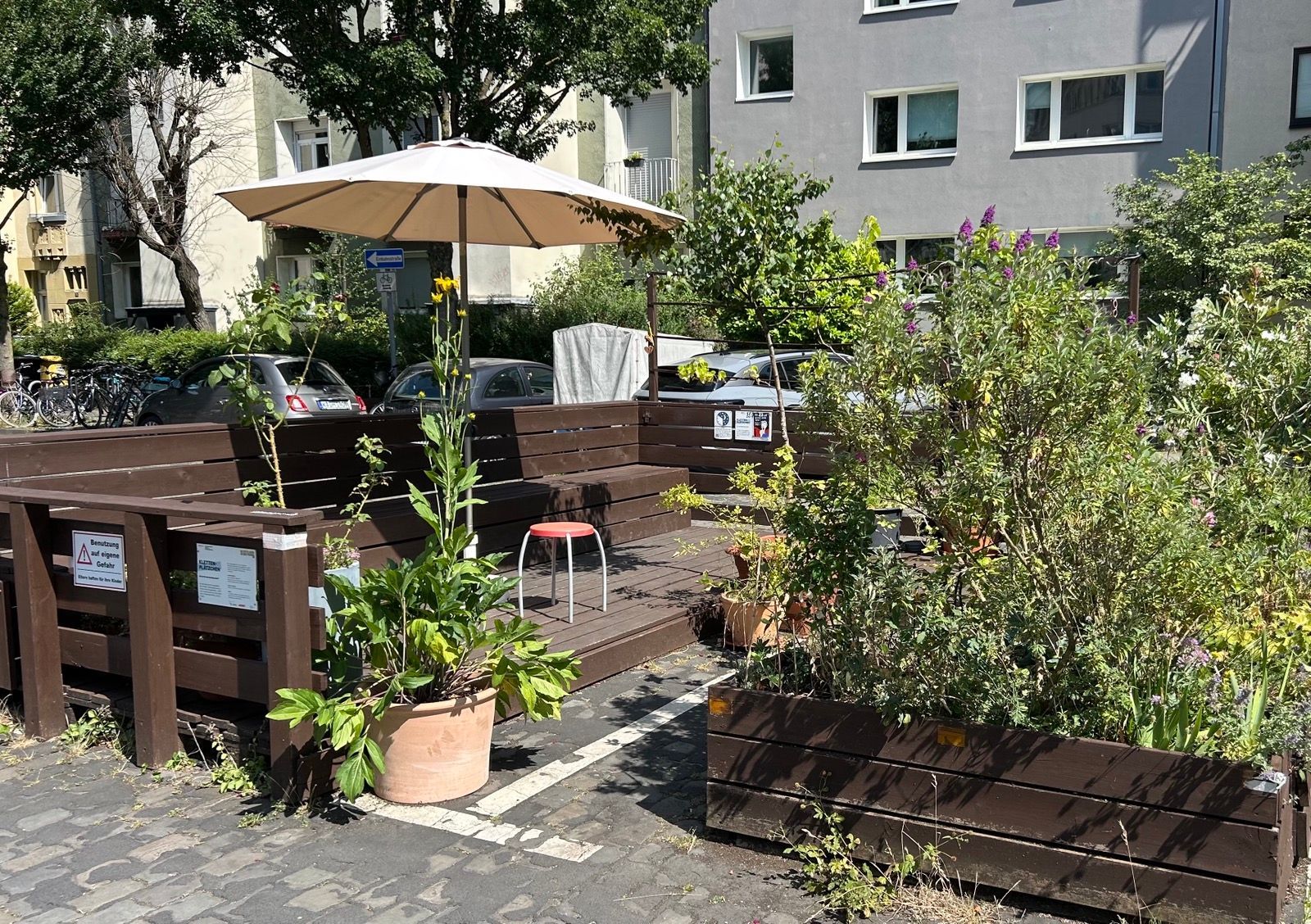 Ein Sonnenschirm und Pflanzen umrahmen Sitzplätze auf einer Straße.