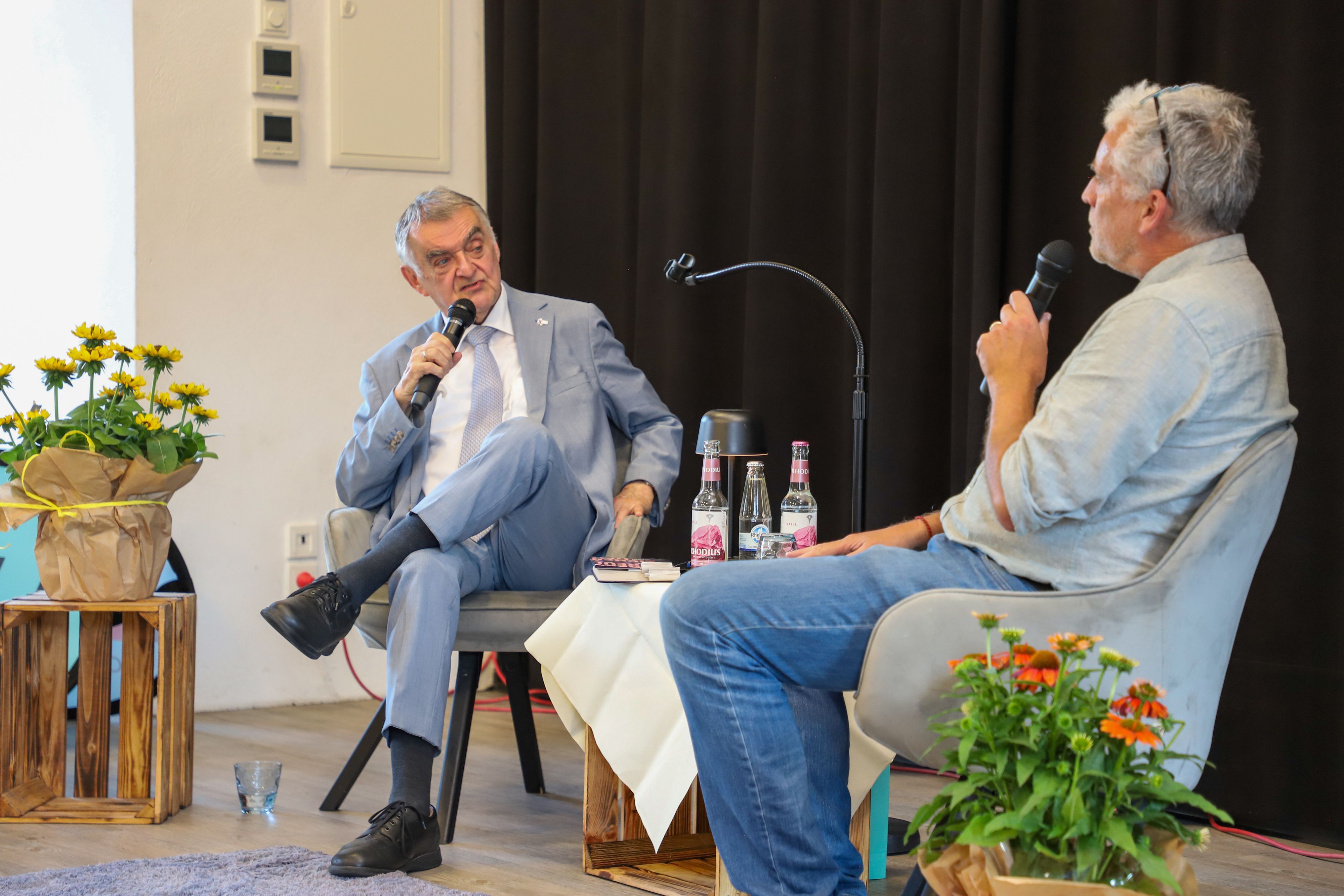 Zwei ältere Männer sitzen auf einer Bühne und sprechen miteinander.