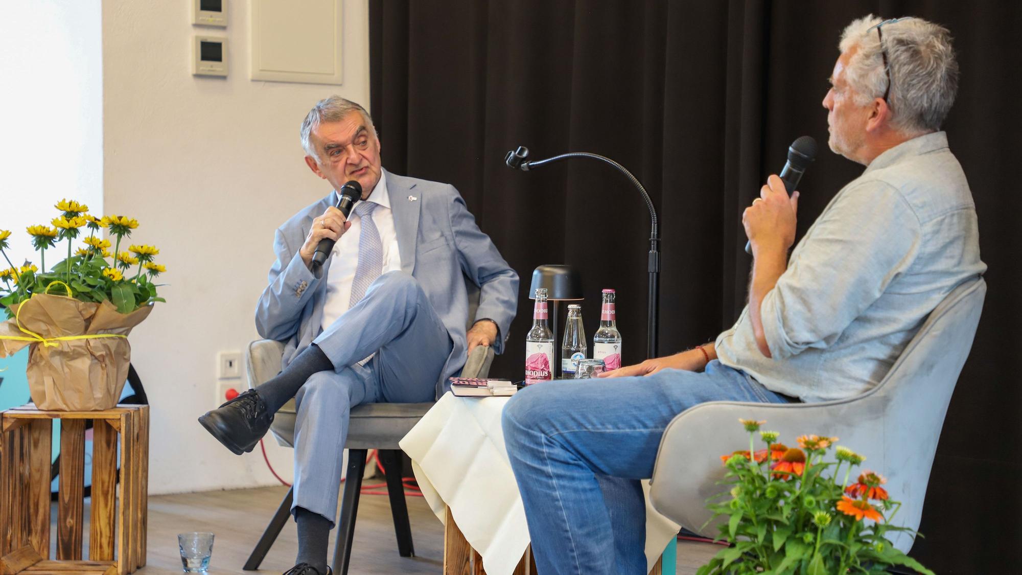 Zwei ältere Männer sitzen auf einer Bühne und sprechen miteinander.