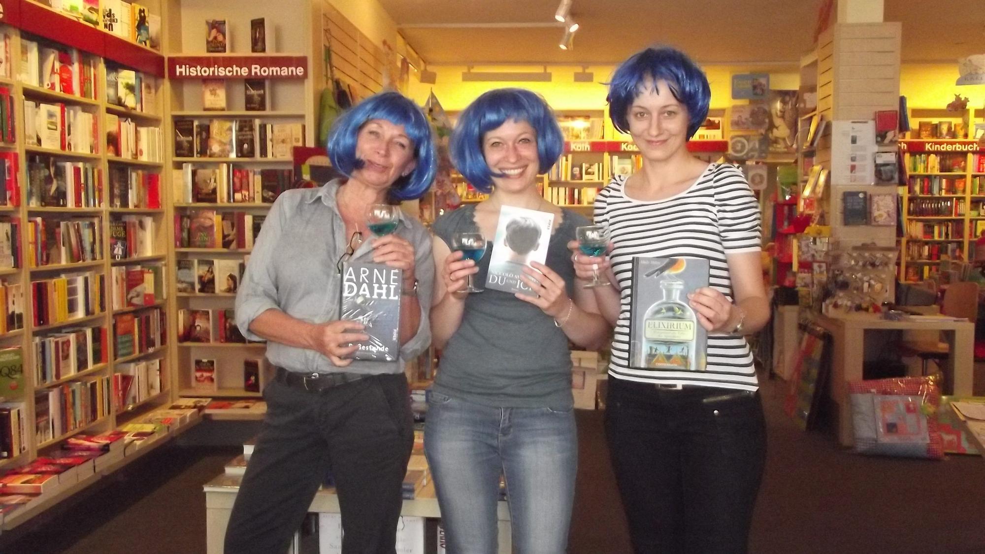 Drei Frauen mit blauen Perücken stehen in einer Buchhandlung nebeneinander.