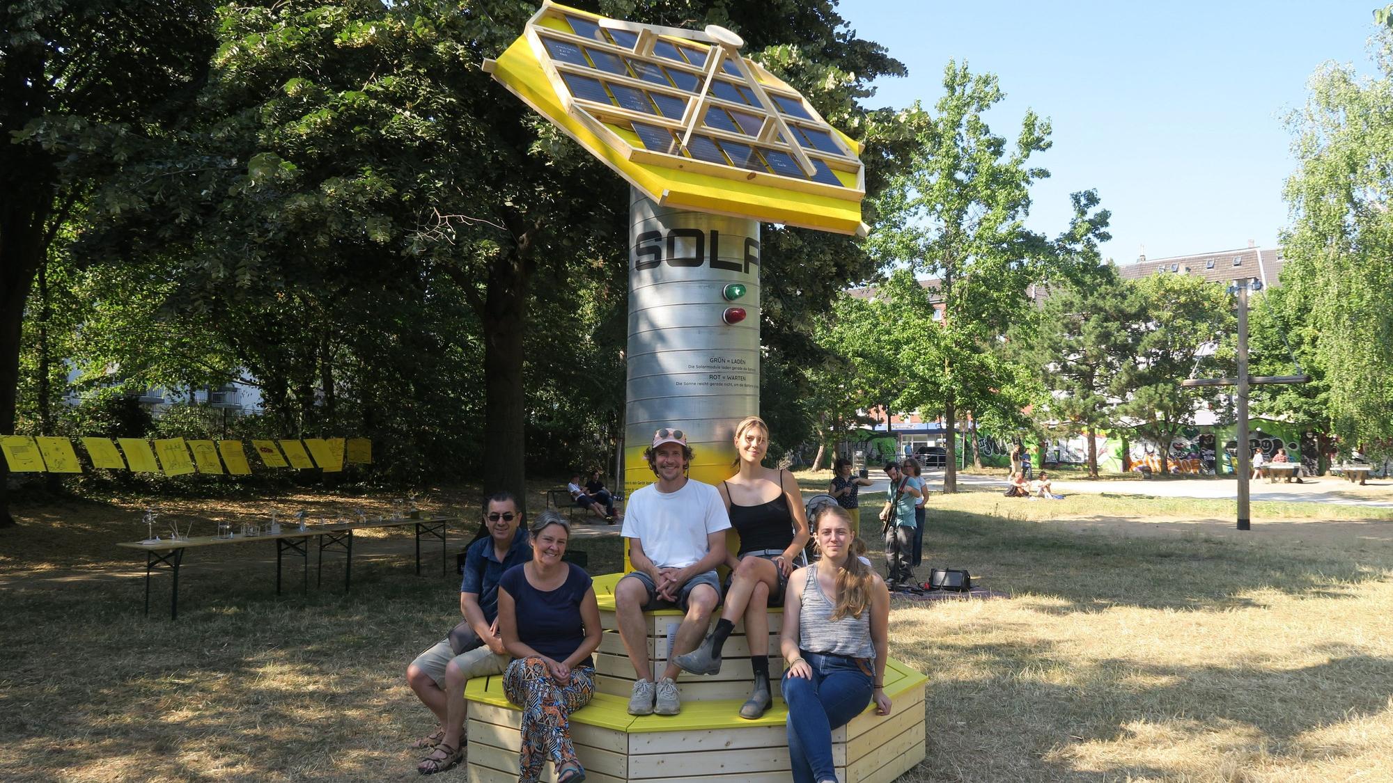 Fünf Menschen sitzen auf Sitzbänken, die eine Solarskulptur umgeben.