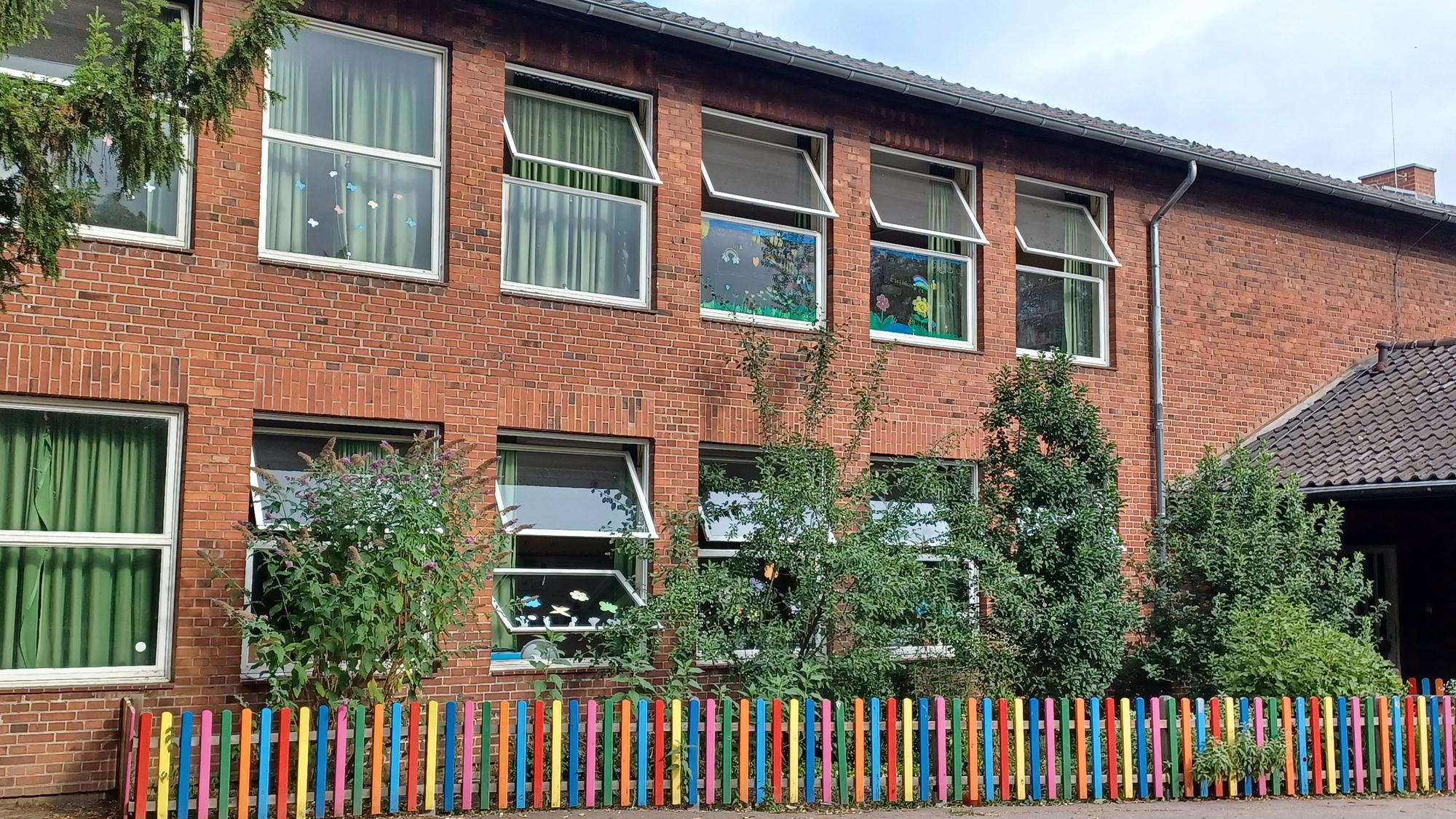 Hinter einem bunten Zaun liegt ein zweigeschossiges Schulgebäude mit großen Fenstern.