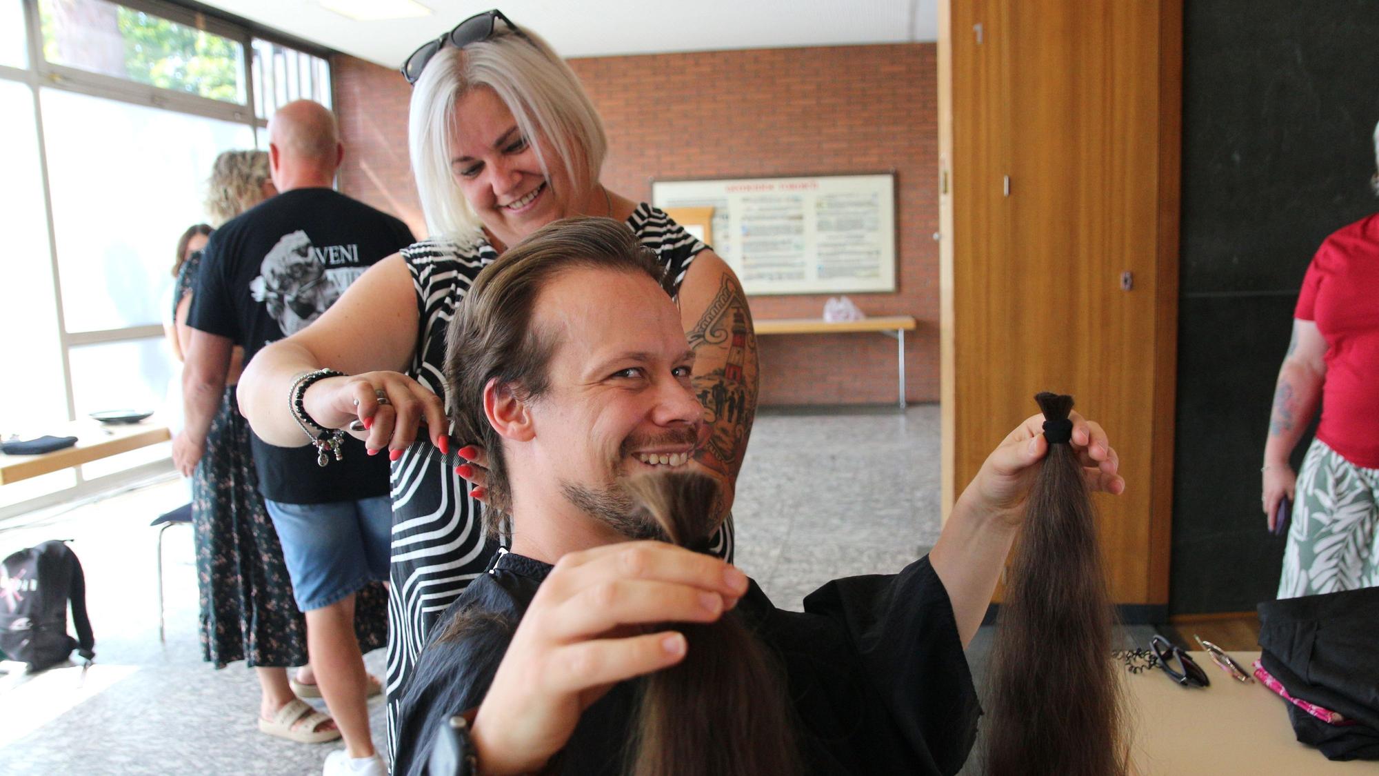 Das Bild zeigt, wie ein Mann seinen abgeschnittene Zöpfe hält und lächelt. Hinter ihm ist die Frisuerin noch an seinen Haaren am Werk.