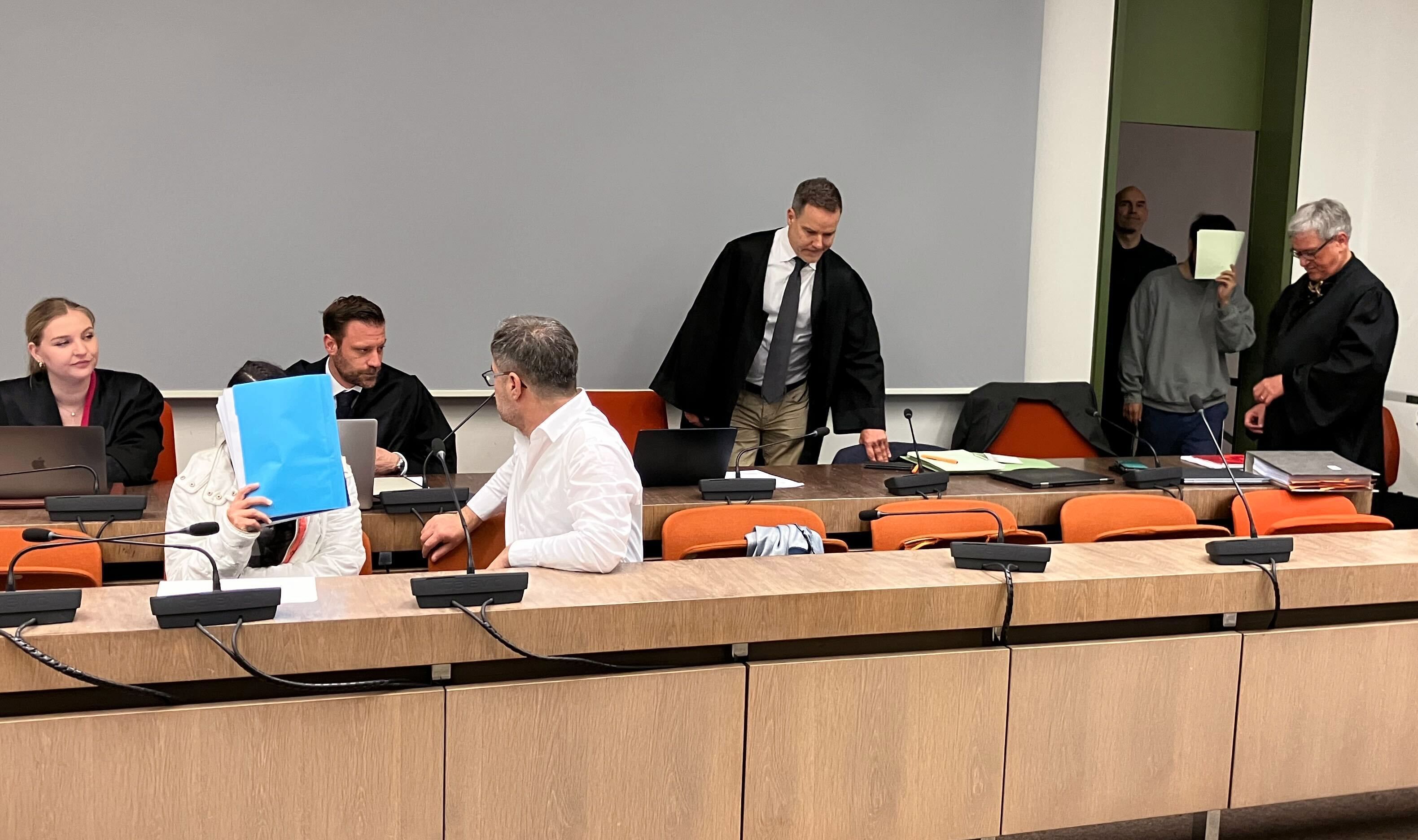 Die wegen Versklavung, Vergewaltigung und Verbrechen gegen die Menschlichkeit angeklagte irakische Frau (2.v.l mit Mappe vor dem Gesicht) sitzt im Saal des Oberlandesgerichts, während ihr mitangeklagter Mann (hinten, 2.v.r) ebenfalls in den Saal kommt (Archivbild).