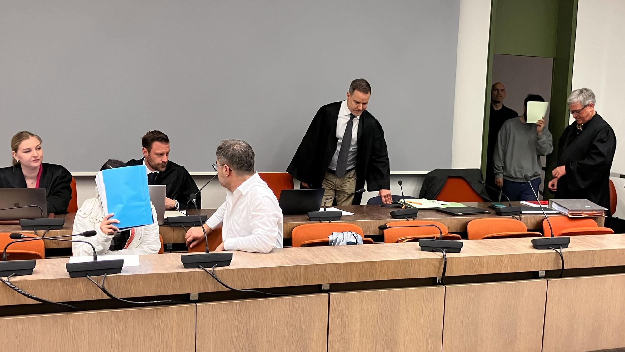 Die wegen Versklavung, Vergewaltigung und Verbrechen gegen die Menschlichkeit angeklagte irakische Frau (2.v.l mit Mappe vor dem Gesicht) sitzt im Saal des Oberlandesgerichts, während ihr mitangeklagter Mann (hinten, 2.v.r) ebenfalls in den Saal kommt (Archivbild).