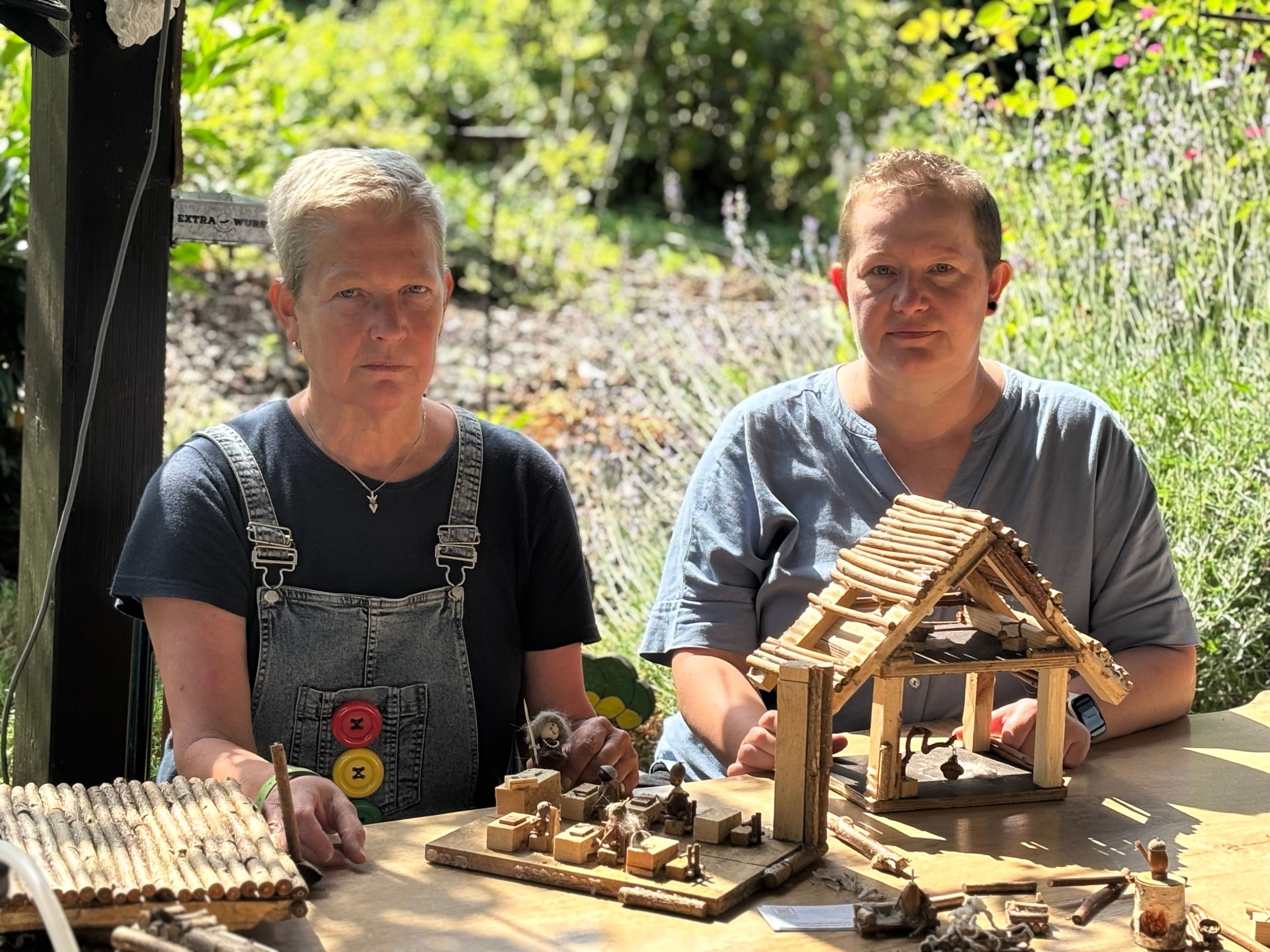 Die Obengenannten sitzen mit versteinerten Gesichtern vor Teilen von Miniatur-Häusern aus Holz, die auf einem Tisch liegen.