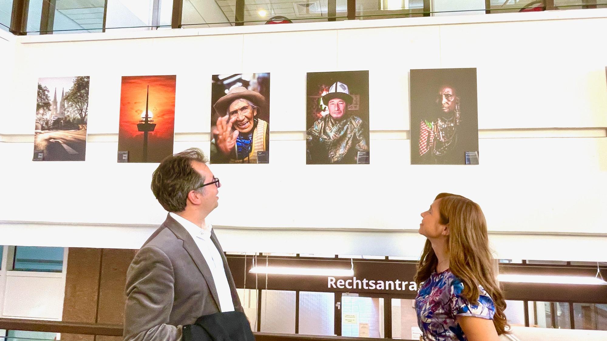 Ein Mann und eine Frau blicken auf eine Wand, an der Bilder im Hochformat hängen.