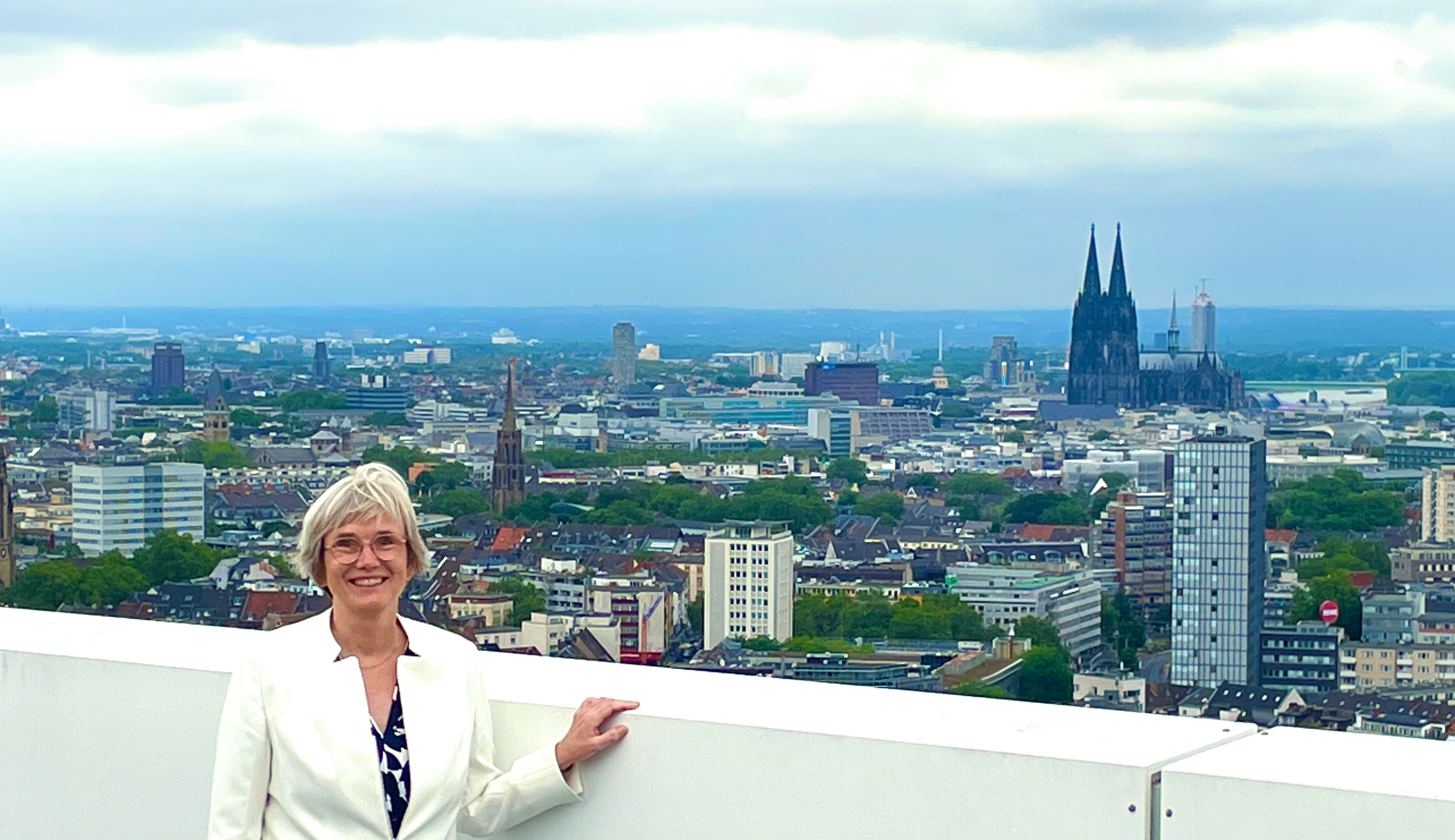 Eine Frau im hellen Blazer mit blondem Haar steht vor einer weißen Brüstung eines Balkons. Hinter ihr ist das Köln-Panorama mit Dom zu sehen.