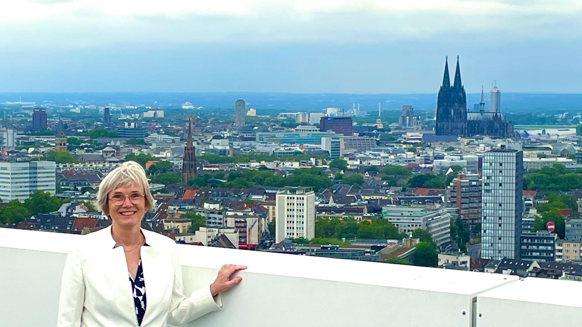 Eine Frau im hellen Blazer mit blondem Haar steht vor einer weißen Brüstung eines Balkons. Hinter ihr ist das Köln-Panorama mit Dom zu sehen.