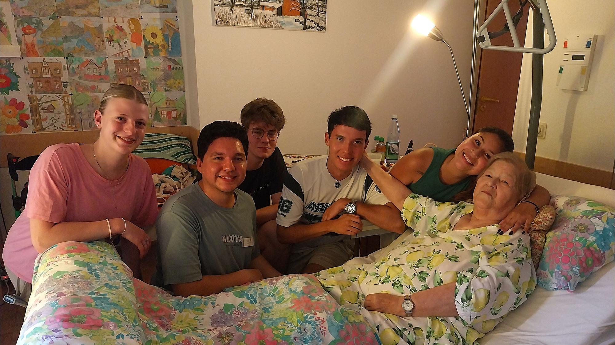 Fünf junge Menschen knien am Bett einer älteren Frau.