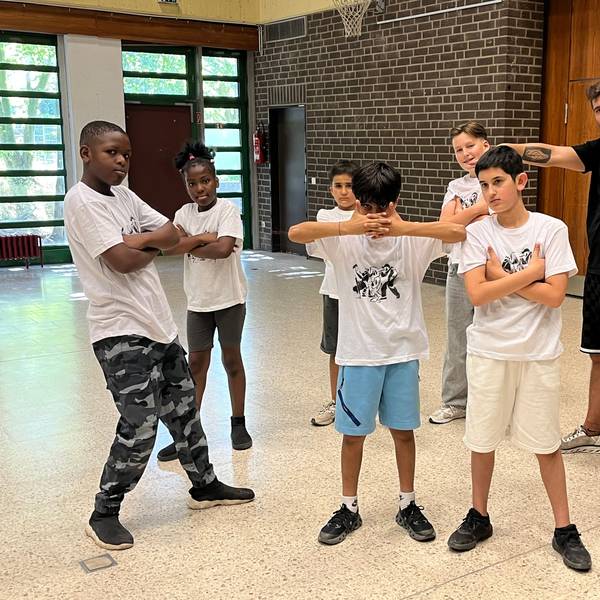 Kinder stehen in einer Turnhalle in coolen Posen. Ein Mann in schwarzem Shirt und schwarzer Hose breitet die Arme aus.