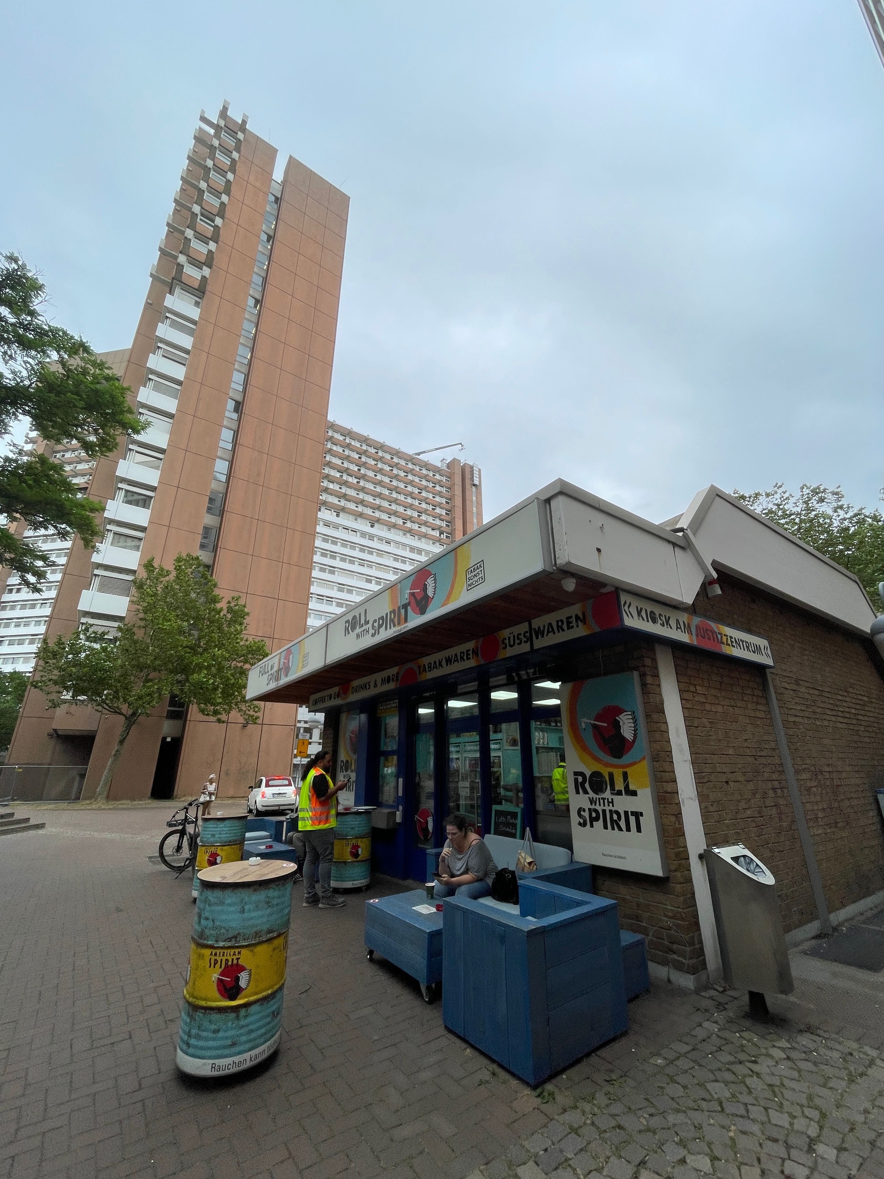 Hinter einem Kiosk, vor dem es eine Sitzecke aus eckigen Holzmöbeln gibt, ragen die Türme des Justizzentrums in die Höhe.