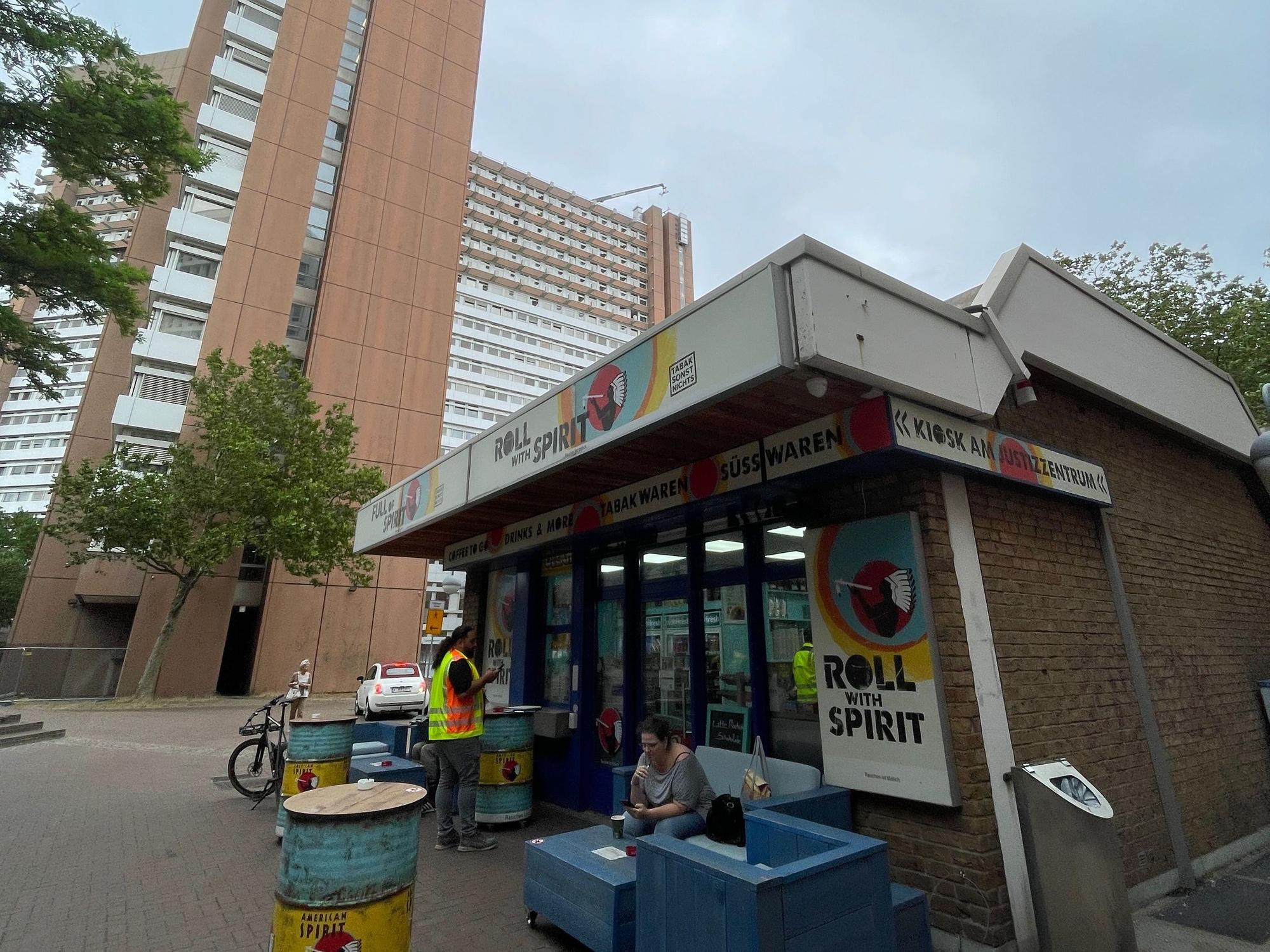 Hinter einem Kiosk, vor dem es eine Sitzecke aus eckigen Holzmöbeln gibt, ragen die Türme des Justizzentrums in die Höhe.