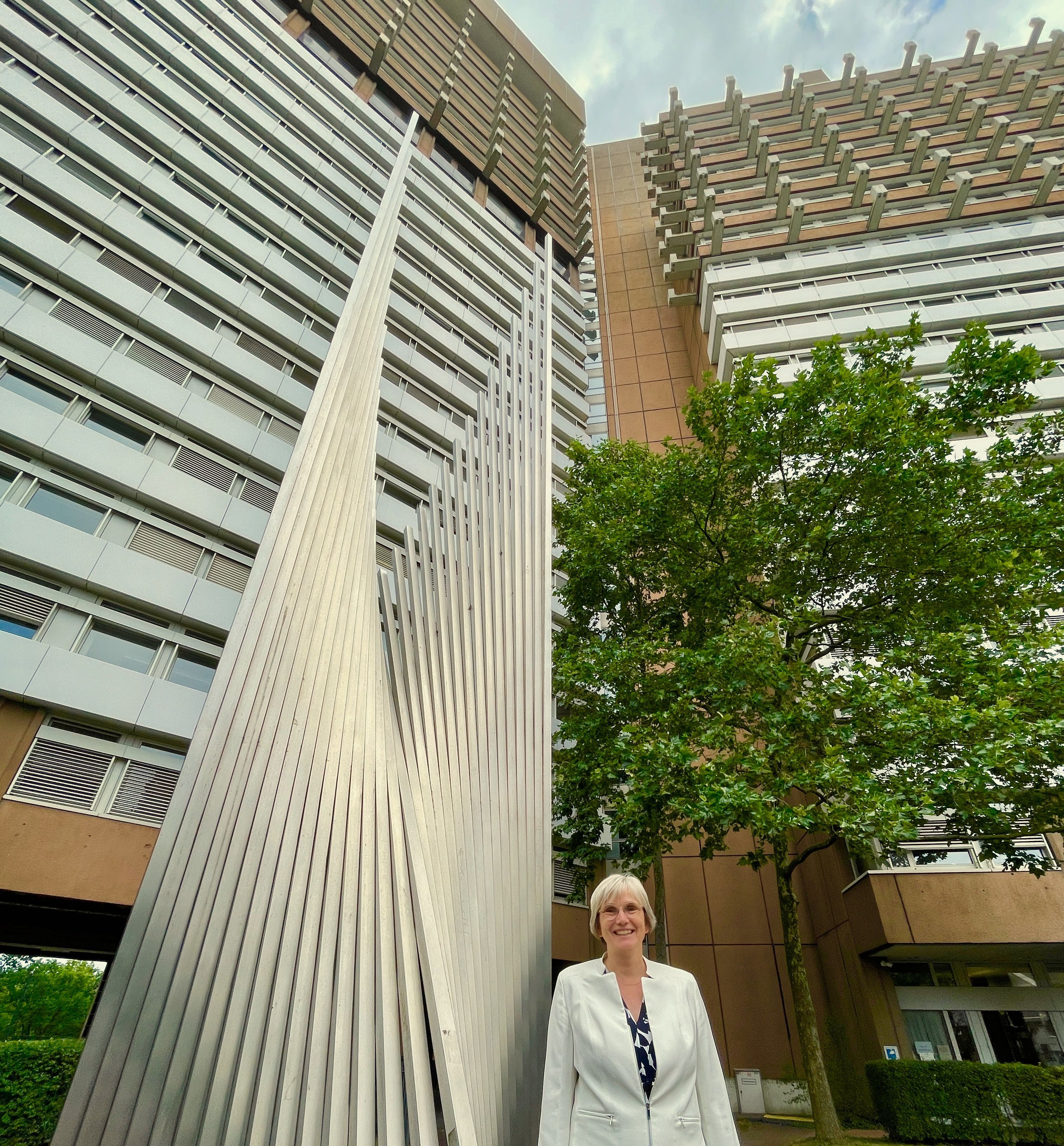 Eine Frau im weißen Blazer mit blonder Kurzhaarfrisur steht neben einer Stahlskulptur vor einem Hochhaus-Komplex.