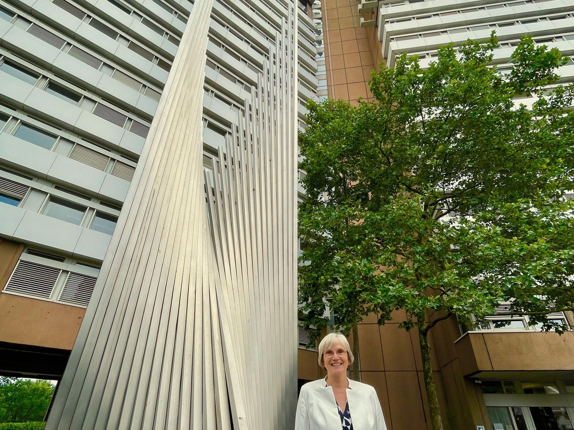 Eine Frau im weißen Blazer mit blonder Kurzhaarfrisur steht neben einer Stahlskulptur vor einem Hochhaus-Komplex.