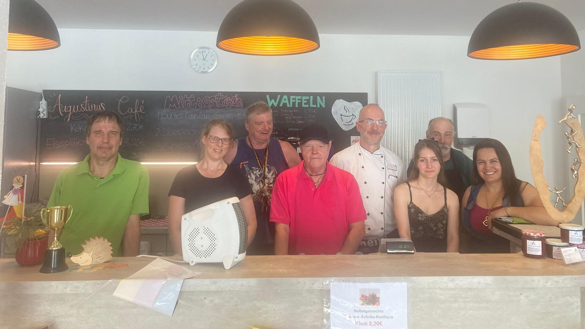 Acht Personen stehen in einem Café an einer Theke, hinter ihnen eine Tafel.