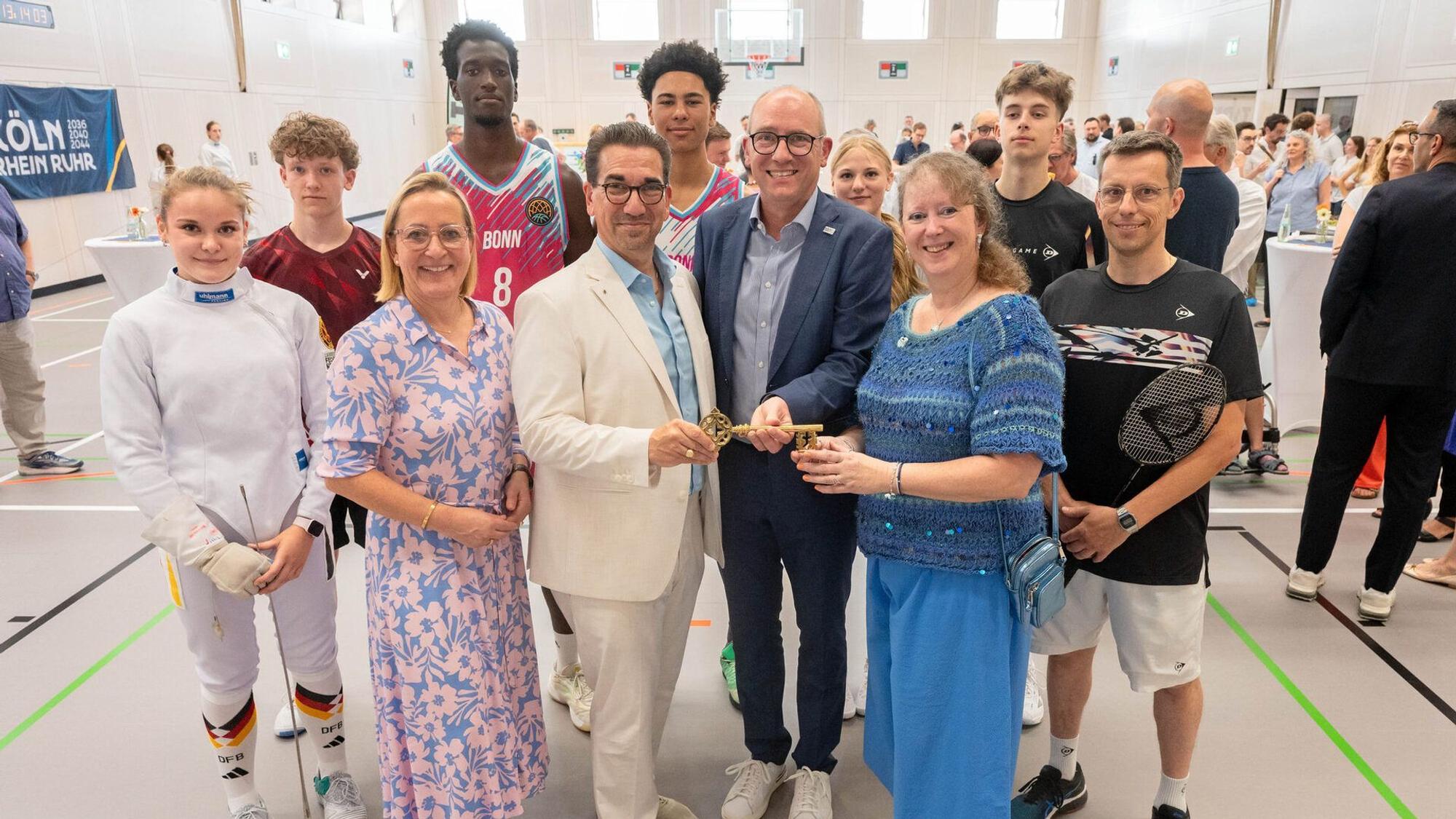 Symbolische Schlüsselübergabe zur Fertigstellung der neuen Sporthalle am Tannenbusch-Gymnasium - vordere Reihe v.l.: Bezirksbürgermeisterin Nicole Bonnie, OB Guido Déus, Schulleiter Eike Schulz und Staatssekretärin Andrea Milz. Hinten v.l.: die Schüler*innen Chiara Rott, Tom Klein, Noah Kouame, Marvin Nduche, Eva Heiden, Jari Hannes sowie Badminton-Trainer Marc Hannes.
