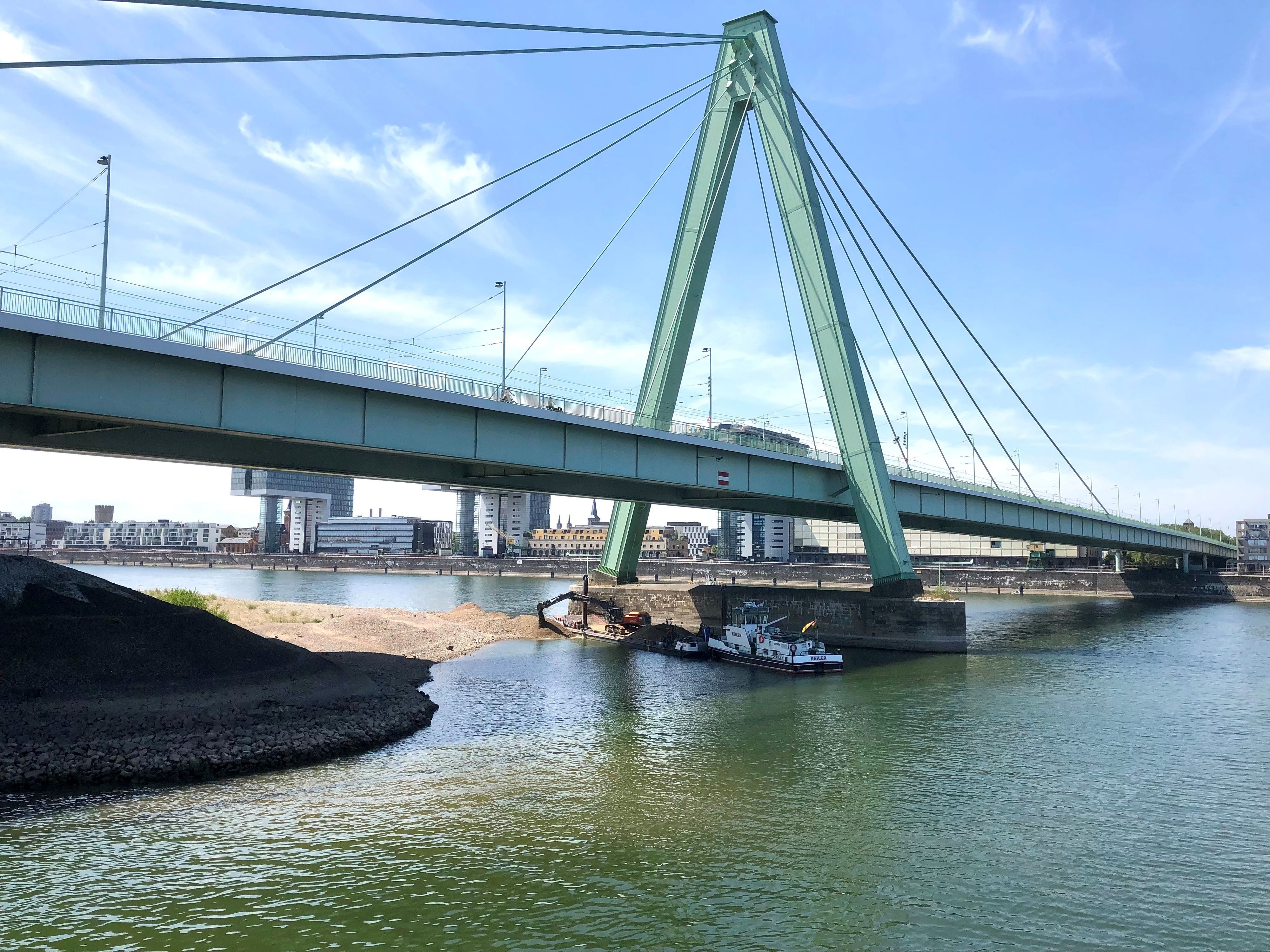 Ein Bagger schüttet Kies aus dem Rhein neben den Pylon der Severinsbrücke. Zurzeit werden in Deutz Untiefen beseitigt.