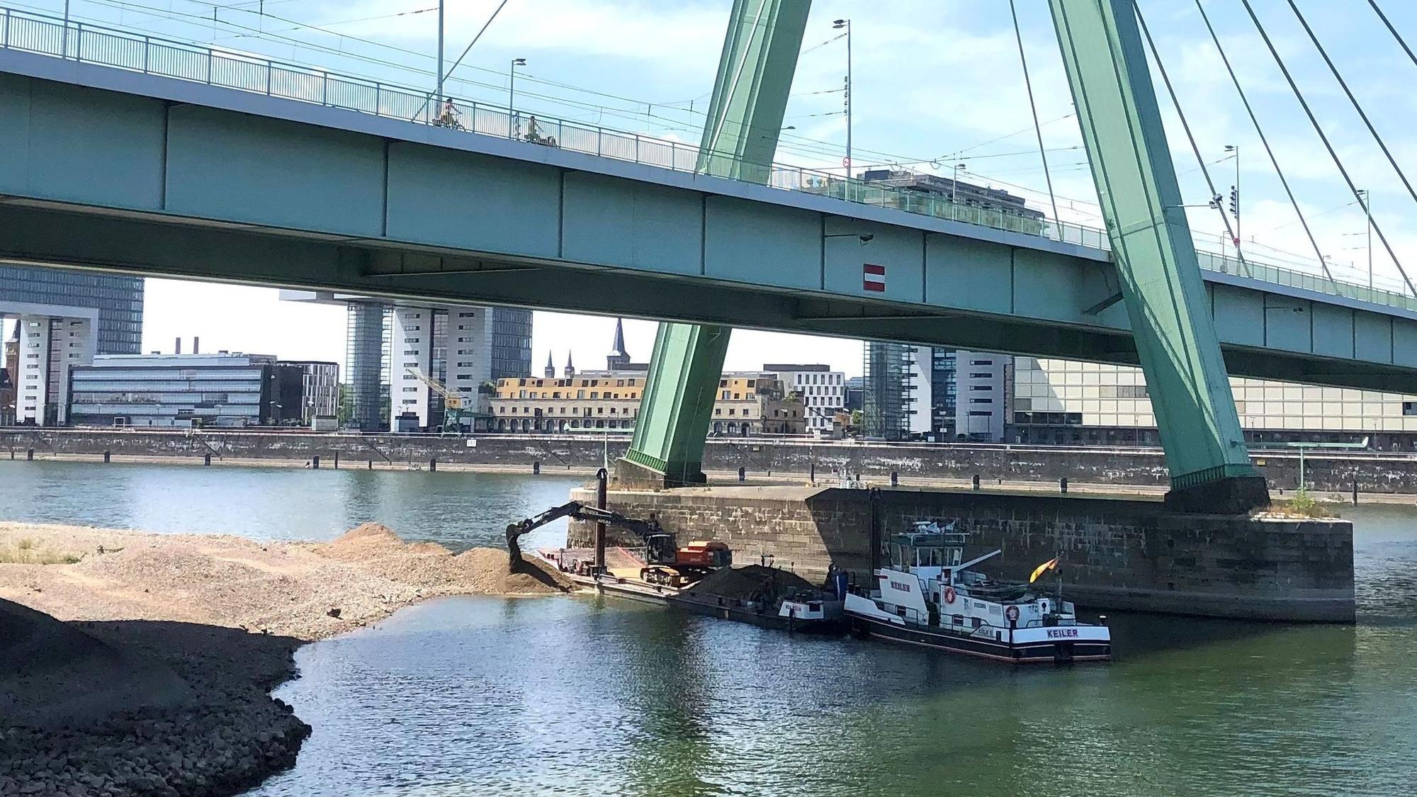 Ein Bagger schüttet Kies aus dem Rhein neben den Pylon der Severinsbrücke. Zurzeit werden in Deutz Untiefen beseitigt.