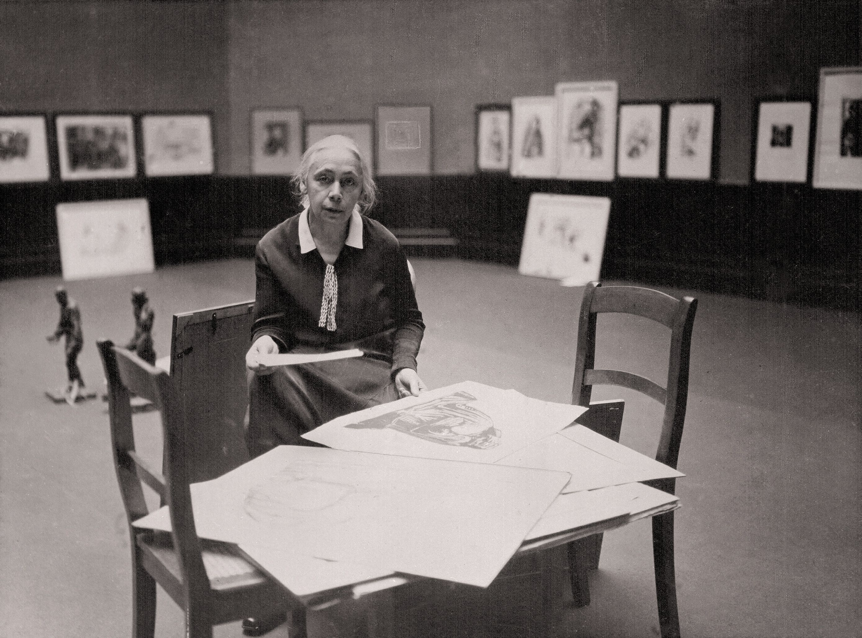 Käthe Kollwitz sitzt in einer Ausstellungshalle.