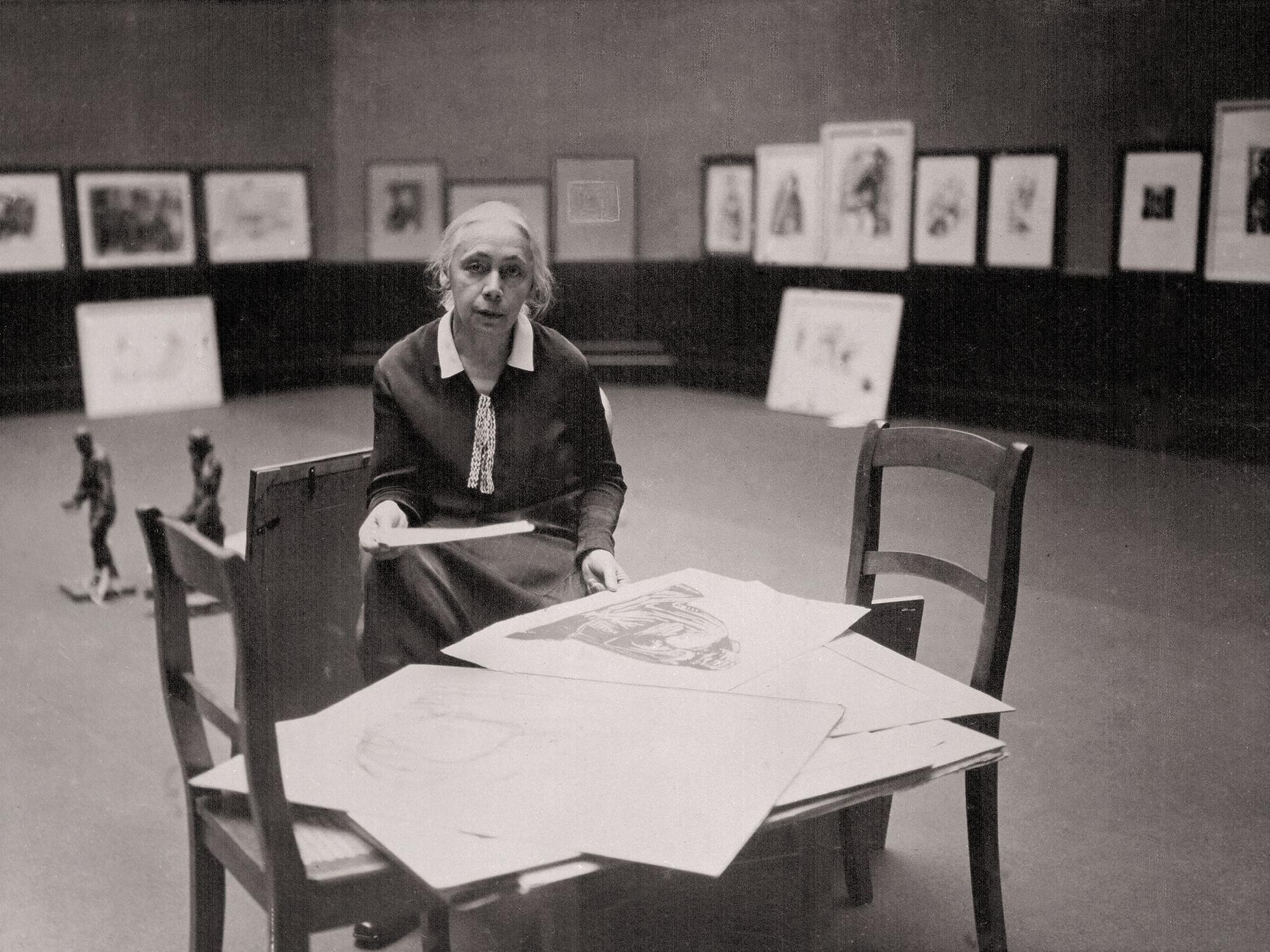 Käthe Kollwitz sitzt in einer Ausstellungshalle.
