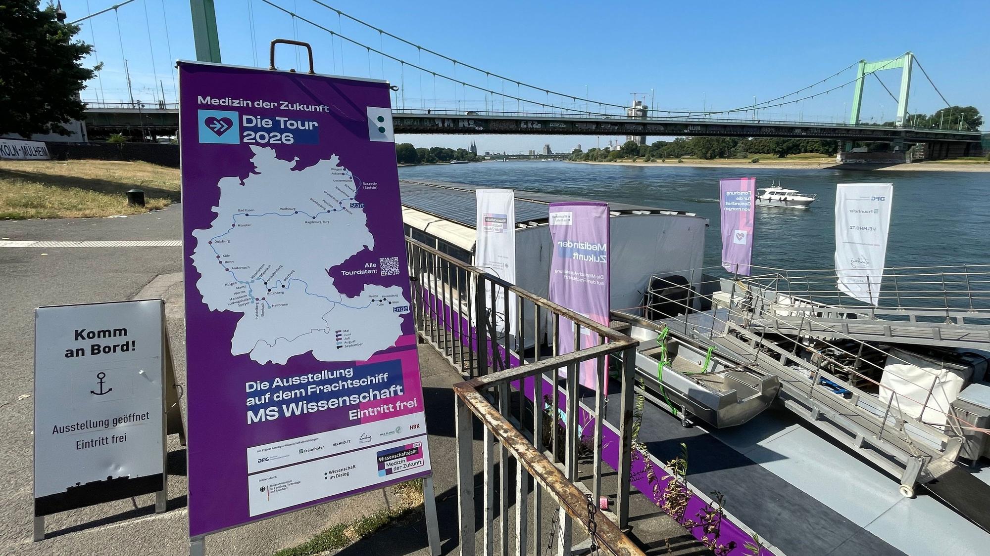 Ein lila Plakat, das die Stationen des Schiffes zeigt, das dahinter am Ufer liegt. Im Hintergrund der Rhein und die Mülheimer Brücke in Köln.