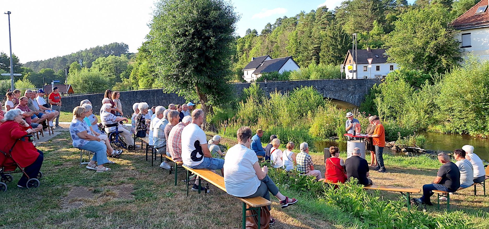 Menschen sitzen auf Holzbänken am Flussufer. Sie richten den Blick auf einen Redner, der an einem Pult steht.