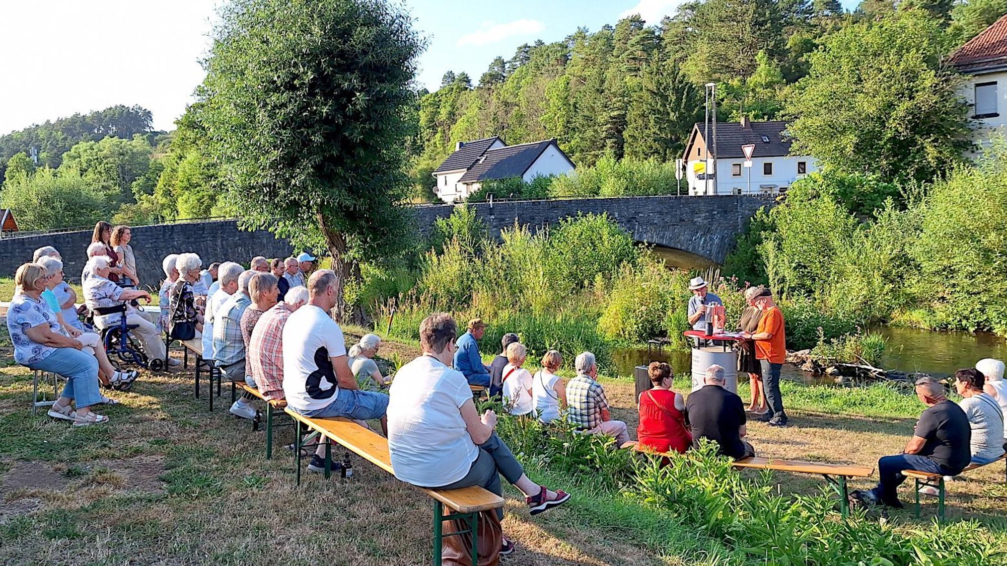 Menschen sitzen auf Holzbänken am Flussufer. Sie richten den Blick auf einen Redner, der an einem Pult steht.