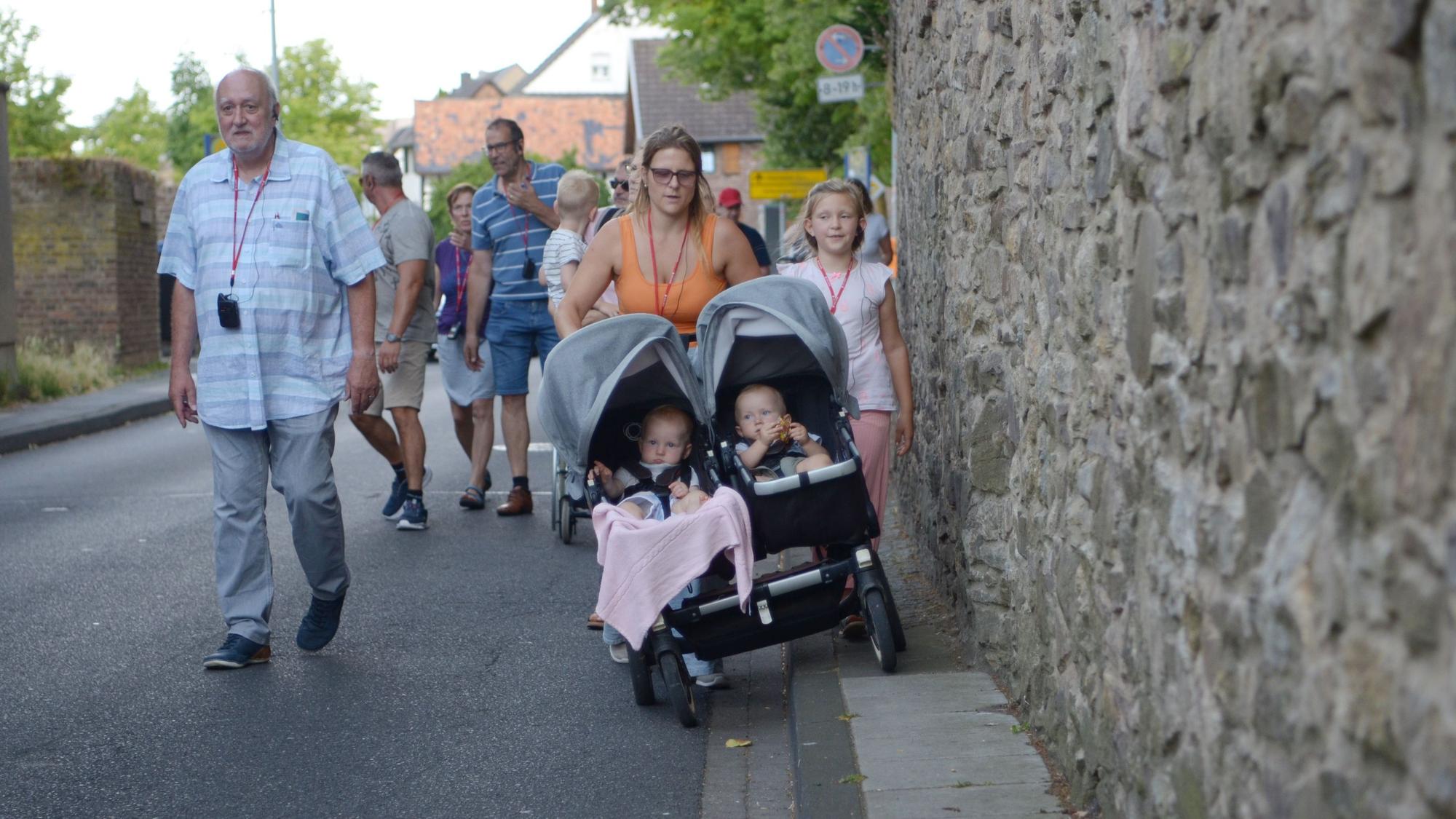 In einer Gruppe von Menschen geht eine Frau, die mit einer Seite ihres Kinderwagens auf die Fahrbahn ausweichen muss, weil der Gehweg zu schmal ist.