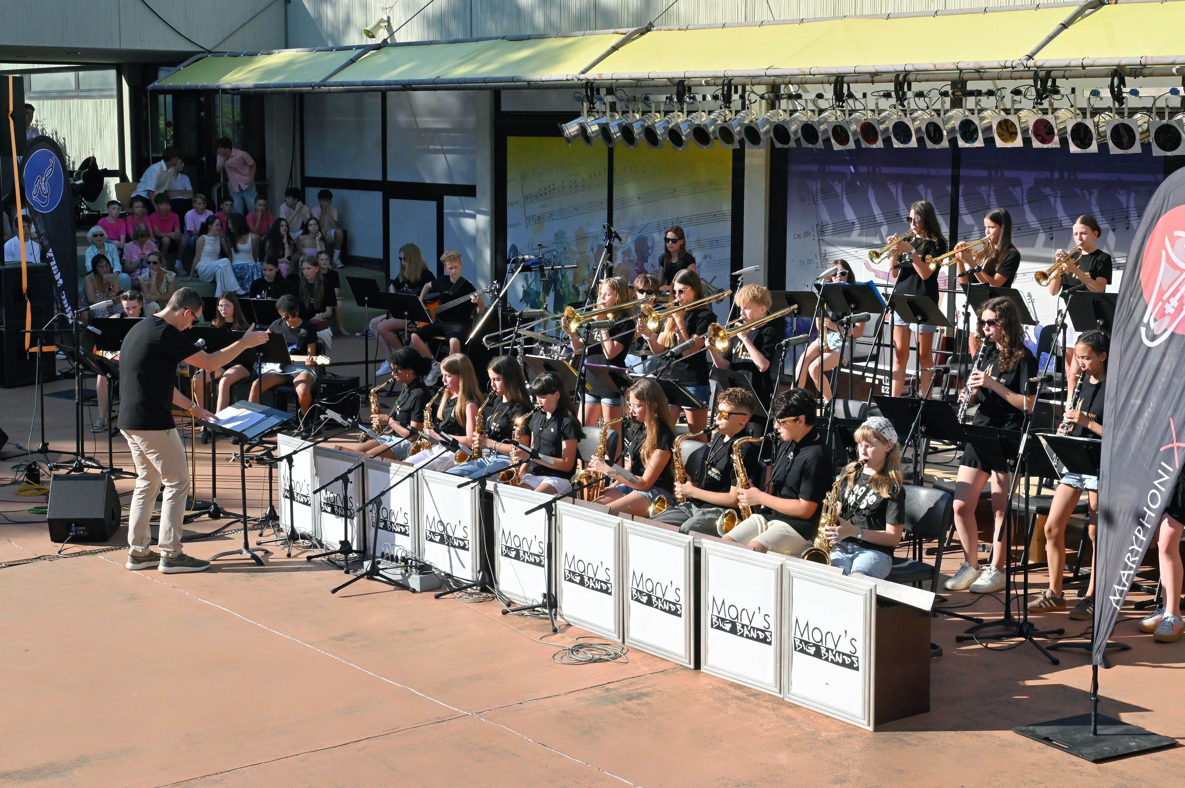Die Musikerinnen und Musiker spielen teils sitzend, teils stehend. Davor steht der Bandleader.