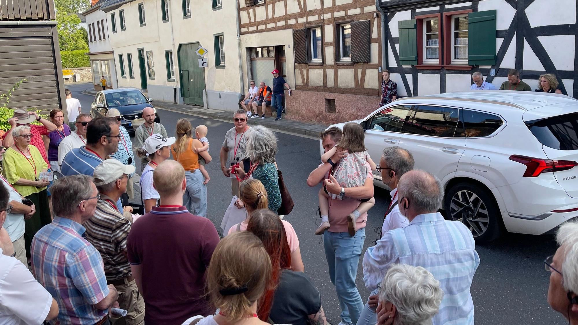 Eine Gruppe von Menschen steht am Straßenrand, Autos fahren an ihnen vorbei.