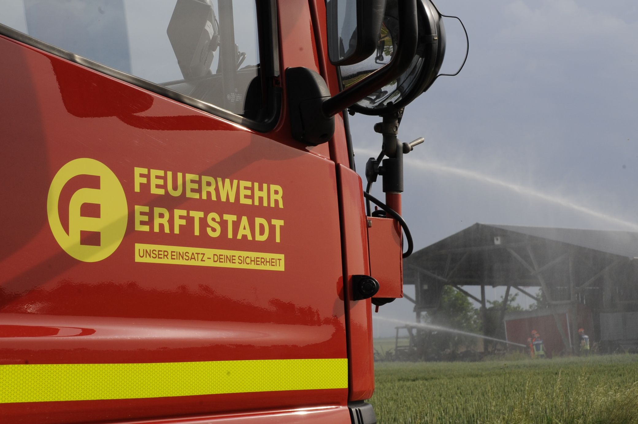 Ein Einsatzfahrzeug der Feuerwehr Erftstadt steht an einem Feld, dahinter sind eine Scheune und Löscharbeiten zu sehen.