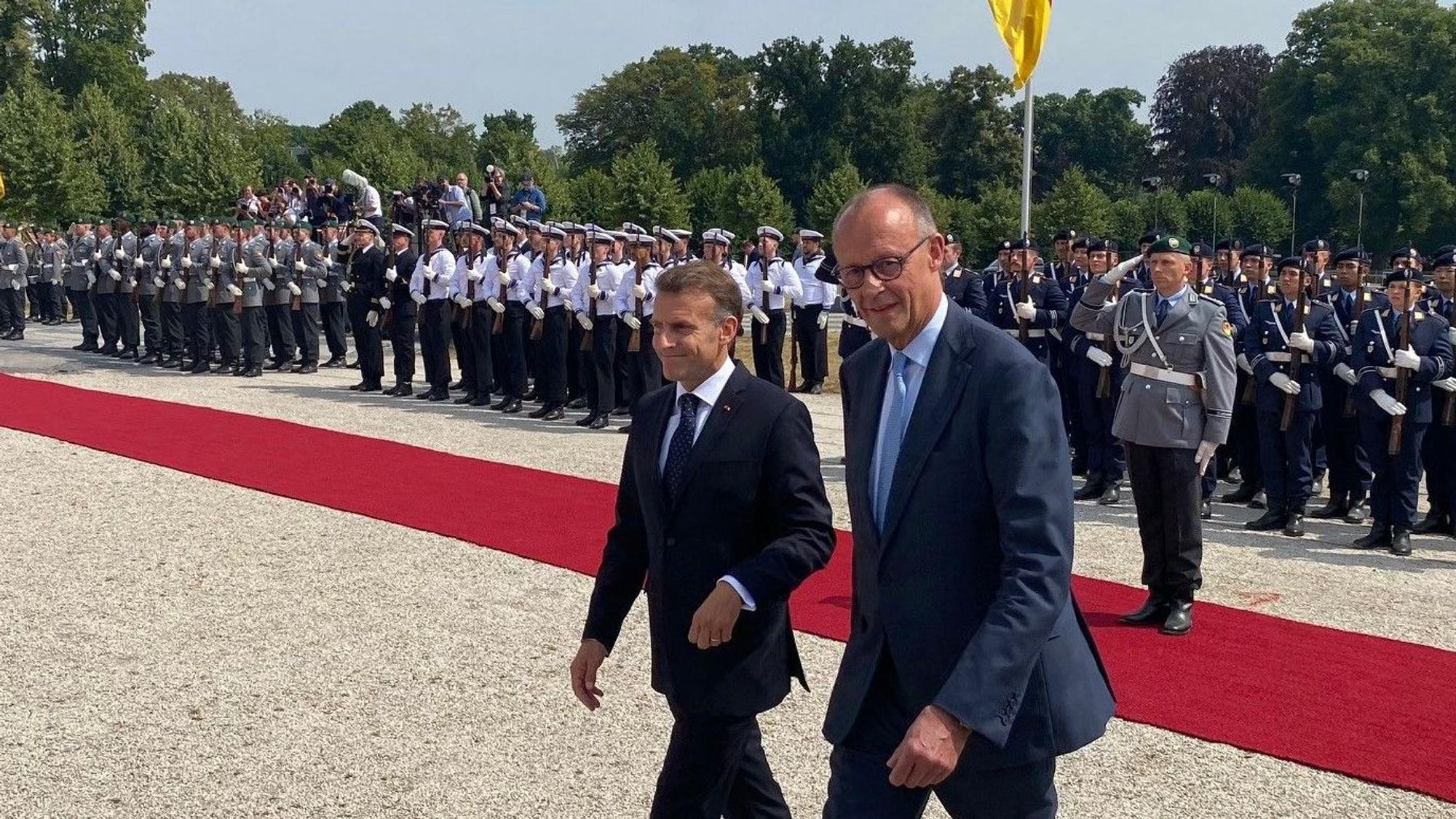Zu sehen sind Bundeskanzler Friedrich Merz und der französische Präsident Emmanuel Macron sowie im Hintergrund Soldaten.