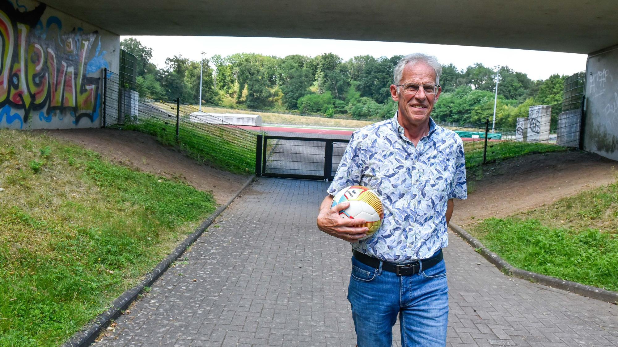 Auf dem Foto ist ein Mann vor einem Stadion zu sehen, er hat einen Fußball in der Hand.