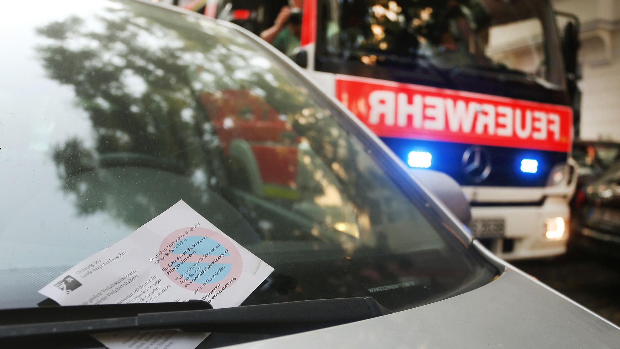 Ein falsch geparktes Auto mit einem Knöllchen, im Hintergrund ist ein Feuerwehrauto mit Blaulicht zu sehen. (Symbolbild)