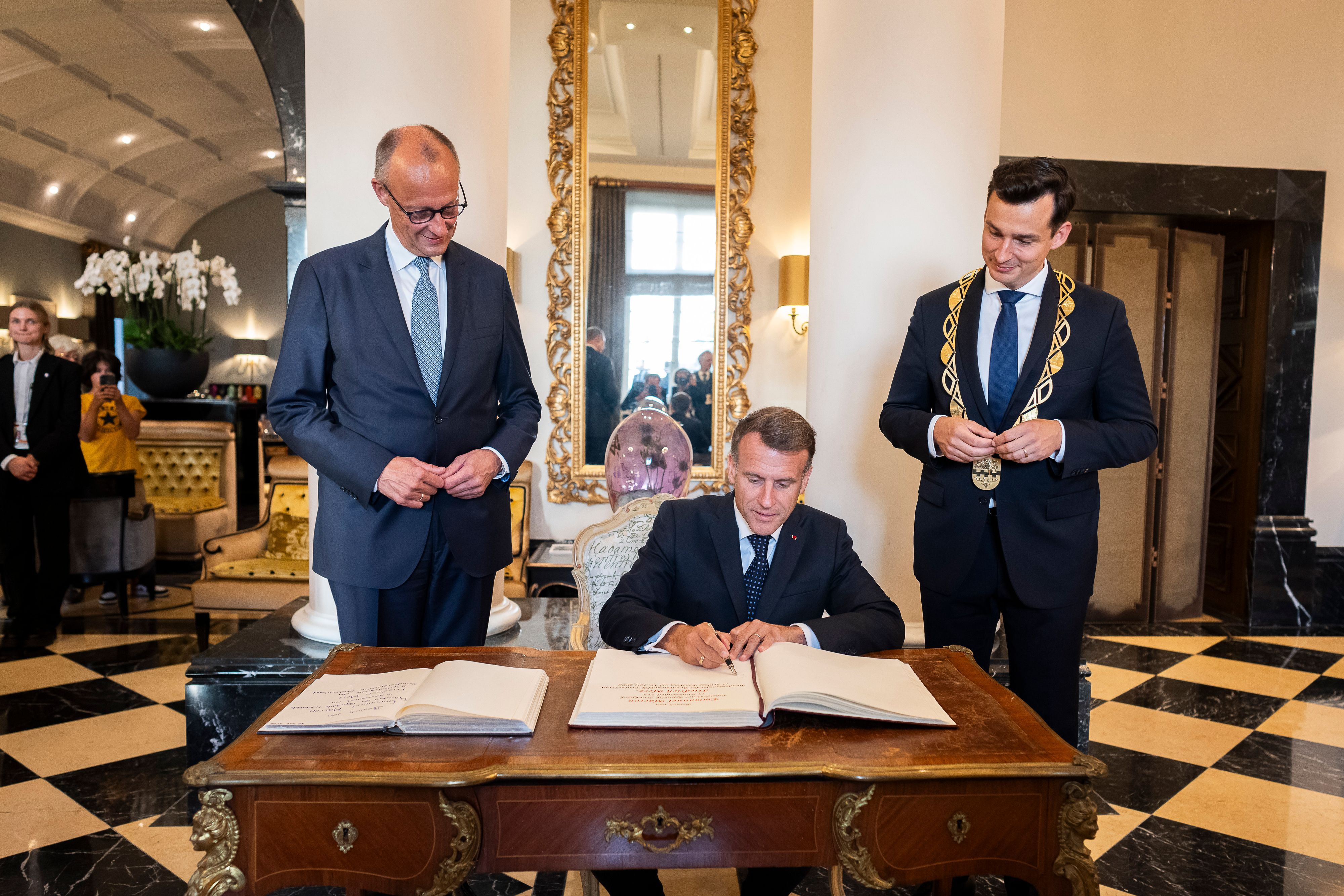 Macron sitzt am Tisch, Merz und der Bürgermeister stehen daneben.