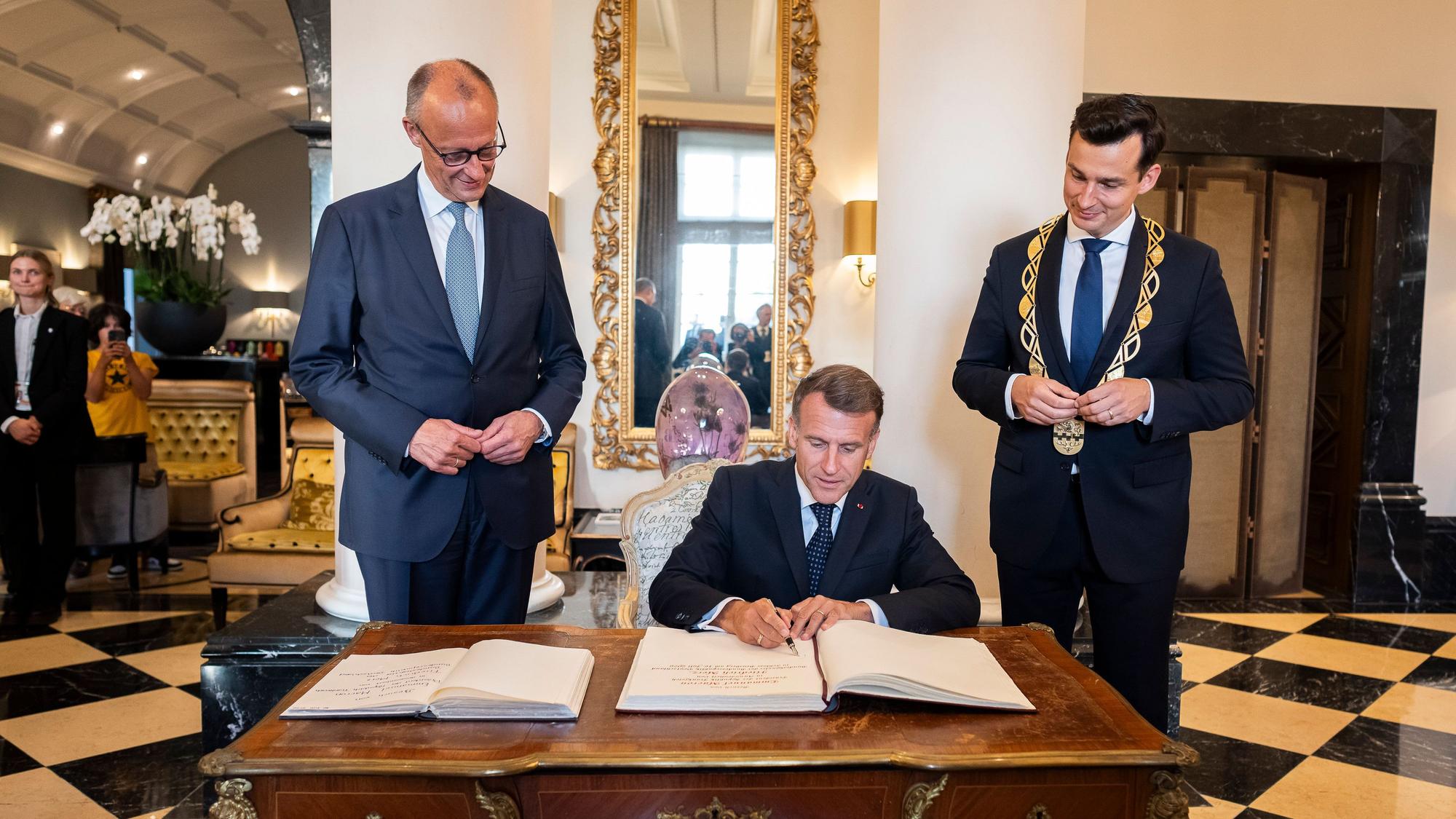 Macron sitzt am Tisch, Merz und der Bürgermeister stehen daneben.