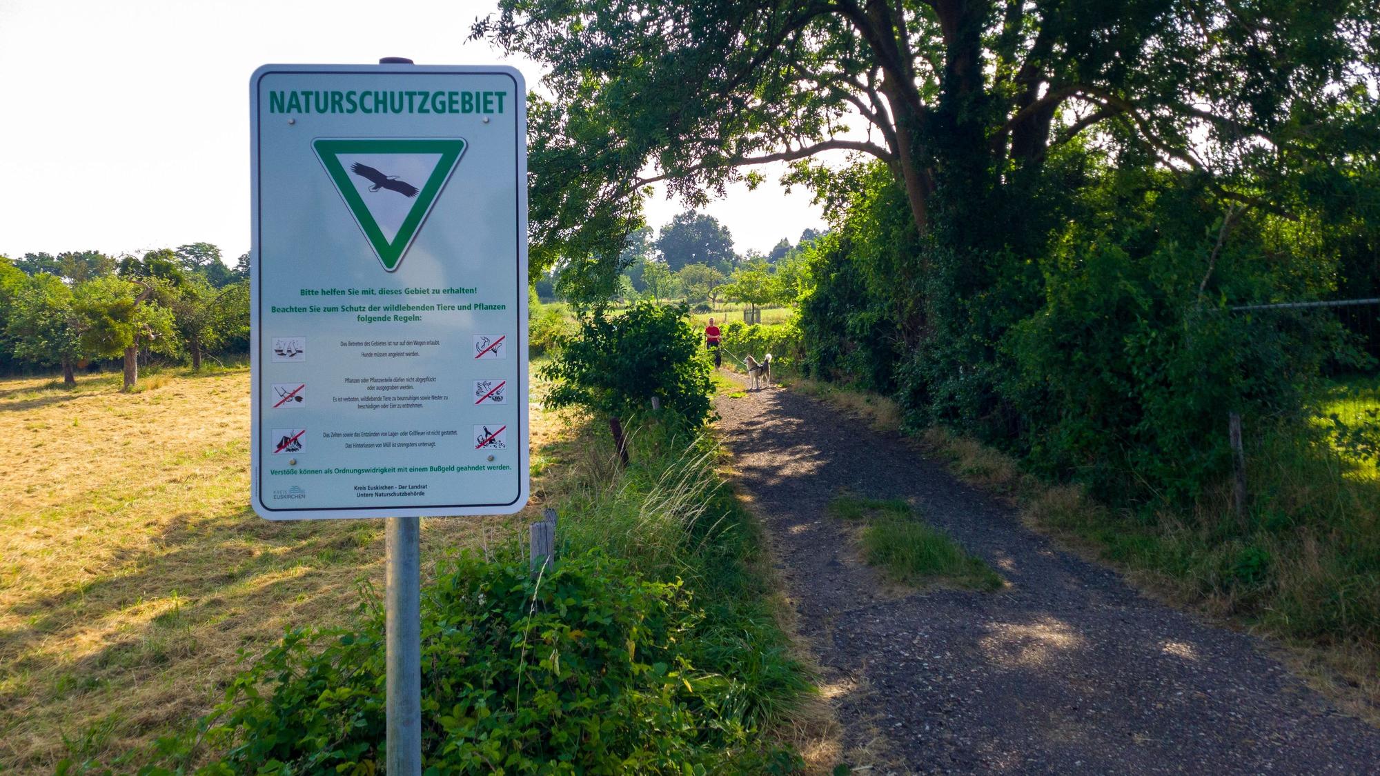 Ein Schild mit der Aufschrift „Naturschutzgebiet“ steht an einem Wanderweg, der an einer Wiese und einem großen Baum entlang führt.