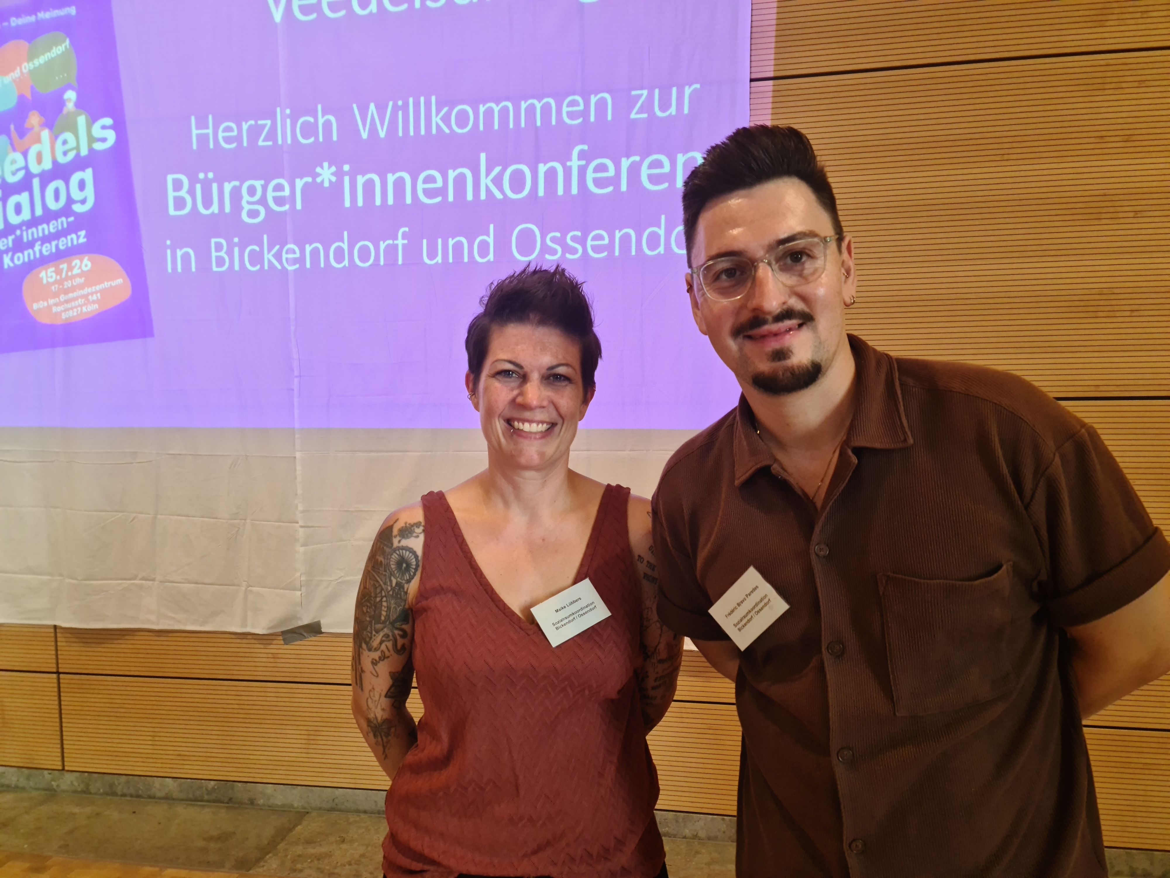 Eine Frau mit kurzem schwarzen Haar und ein Mann mit kurzem schwarzem Haar, Bart und Brille stehen nebeneinander vor einer Beamer-Präsentation, die hinter ihnen zu sehen ist.