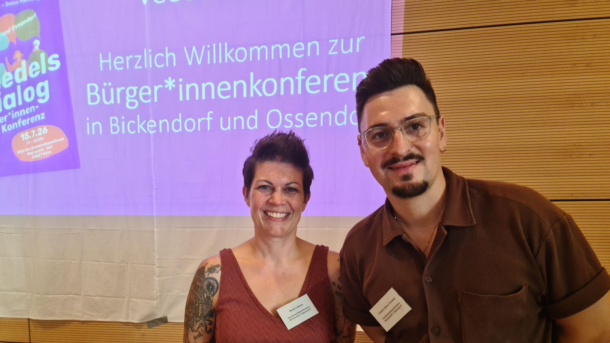 Eine Frau mit kurzem schwarzen Haar und ein Mann mit kurzem schwarzem Haar, Bart und Brille stehen nebeneinander vor einer Beamer-Präsentation, die hinter ihnen zu sehen ist.