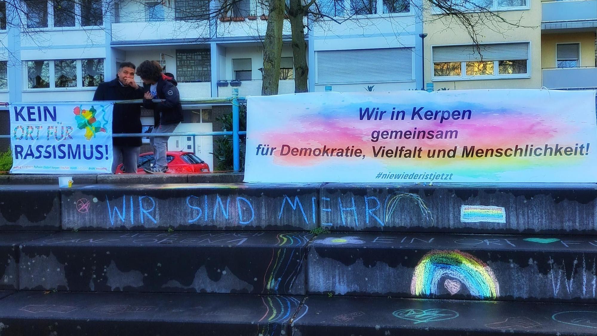 Das Foto zeigt ein Plakat, daneben zwei Menschen.