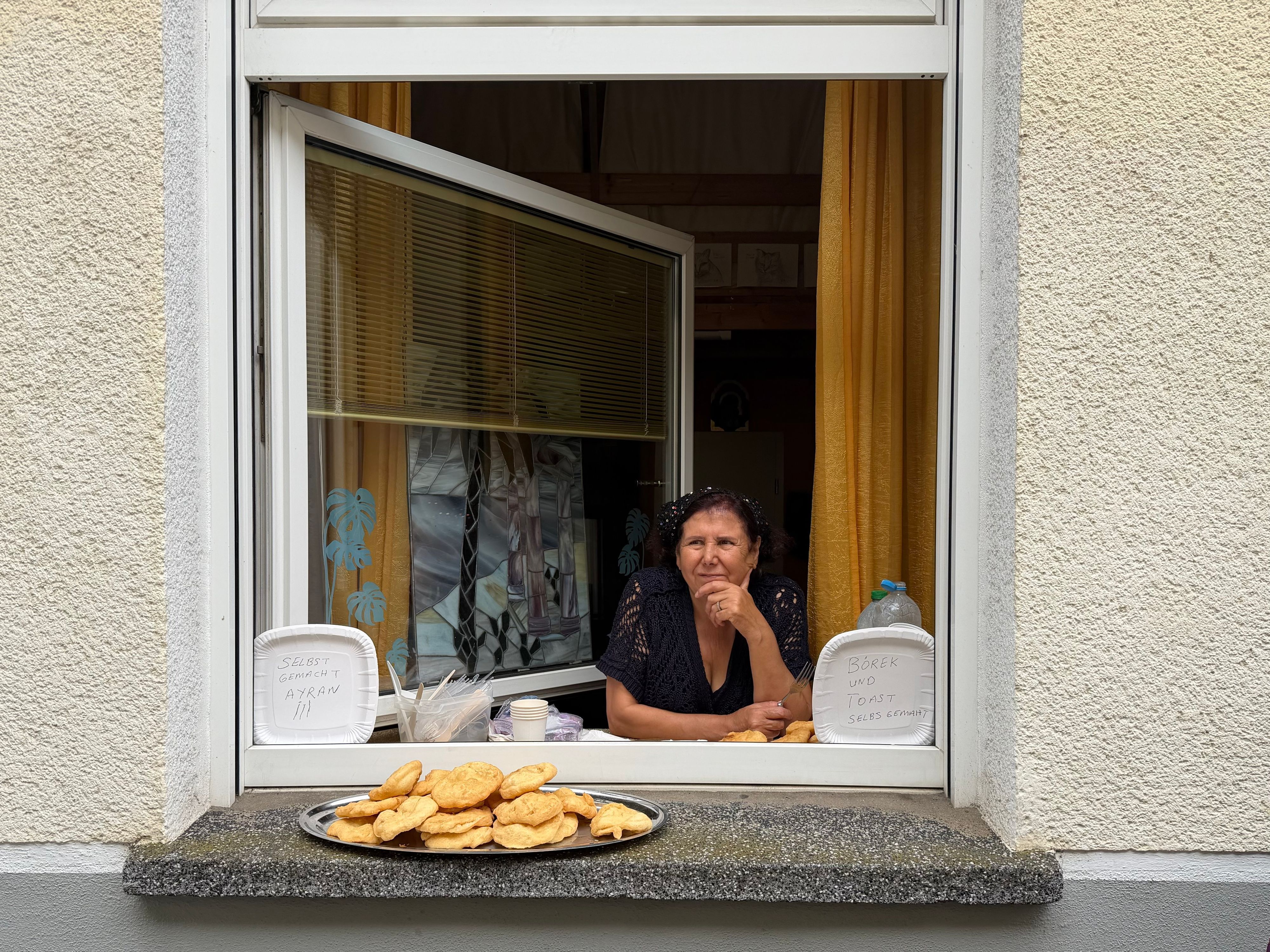 Eine Frau guckt aus ihrem geöffneten Wohnungsfenster, auf dem Fensterbrett steht ein Tablett mit Backwaren.