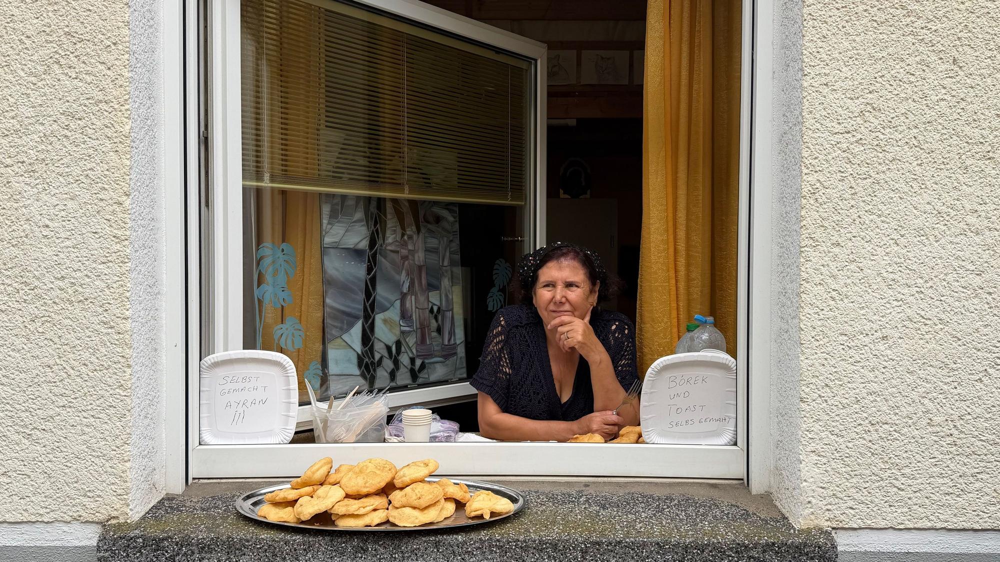 Eine Frau guckt aus ihrem geöffneten Wohnungsfenster, auf dem Fensterbrett steht ein Tablett mit Backwaren.