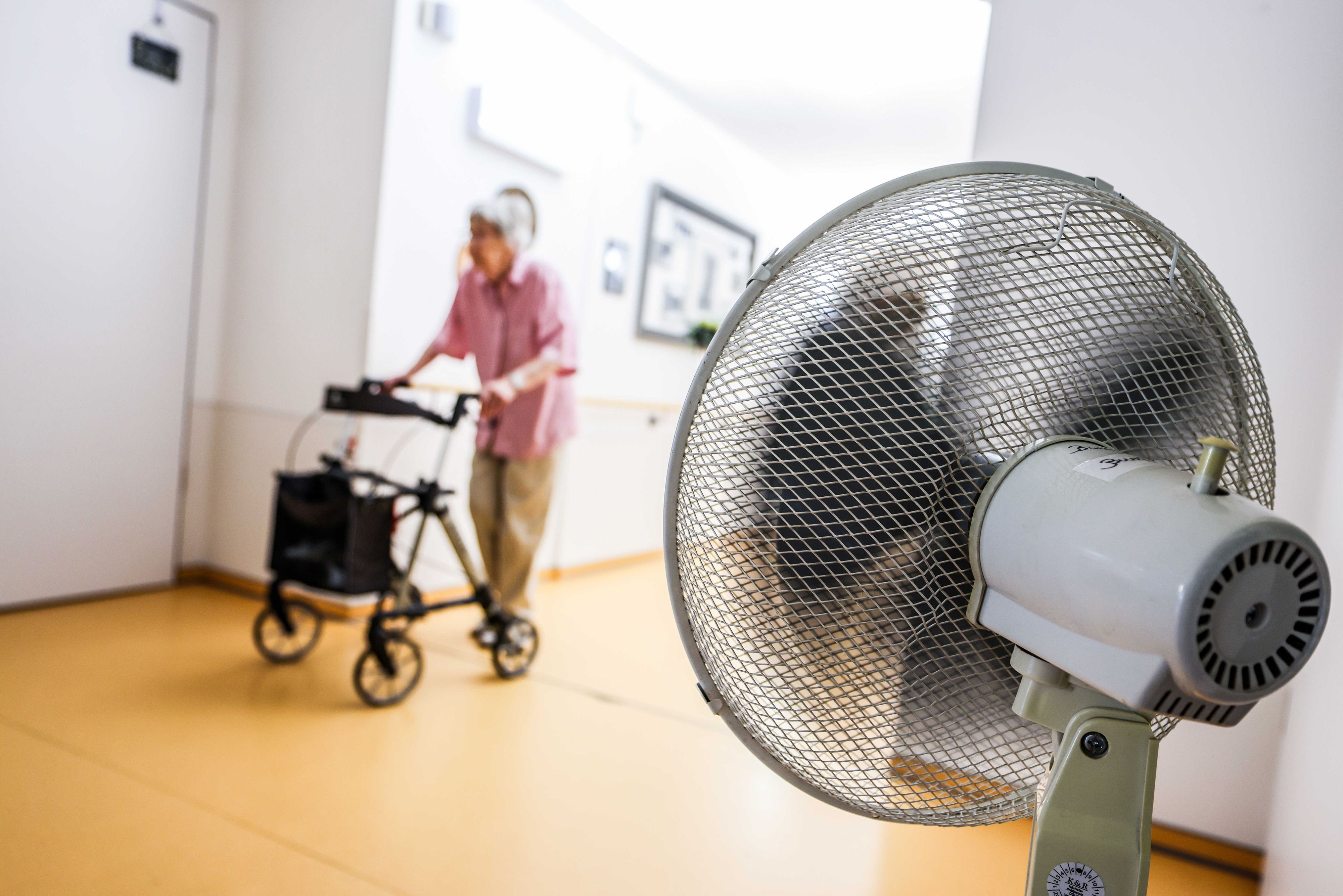 Eine Seniorin schiebt ihren Rollator über einen Flur. Rechts ist im Vordergrund ein Ventilator zu sehen.