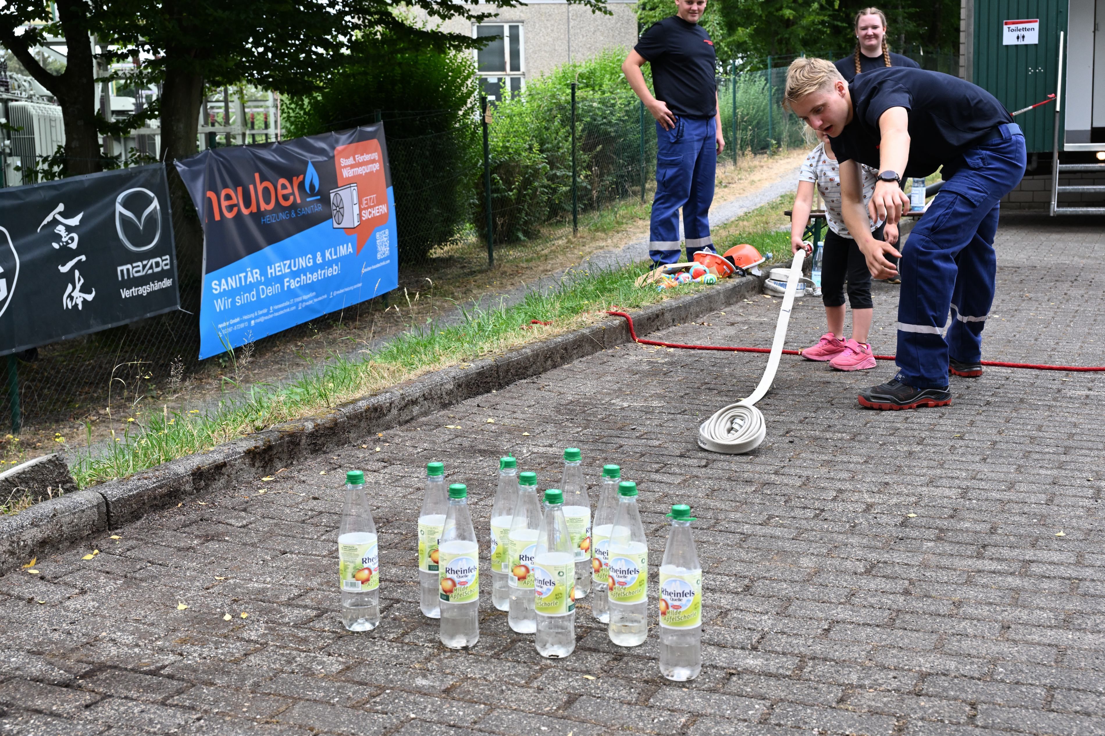 Zu sehen ist ein junger Feuerwehrmann, der einem Kind zeigt, wie man einen Feuerwehrschlauch ausrollt.